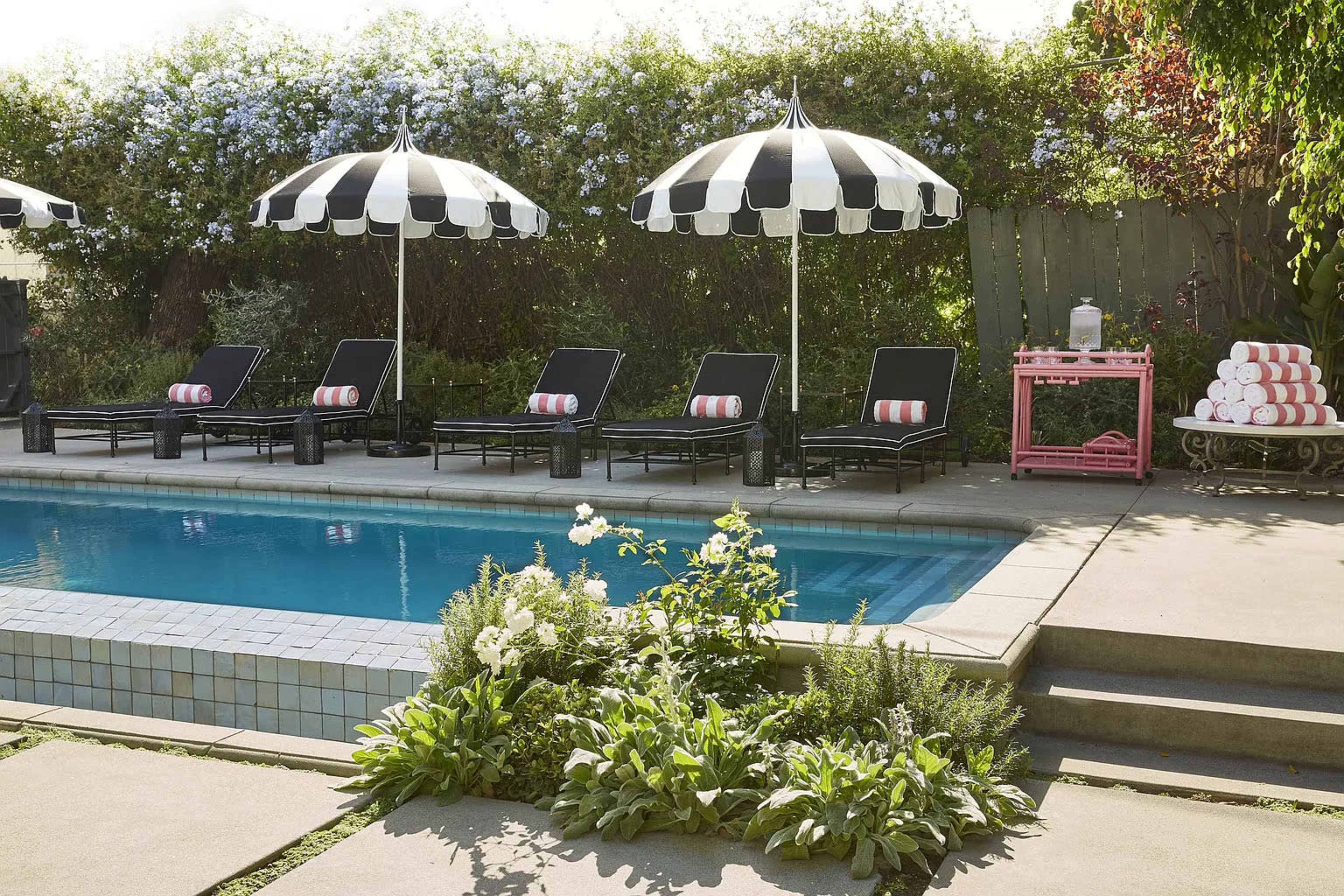 The image shows a swimming pool surrounded by black lounge chairs with striped umbrellas and a service cart with stacked towels nearby.