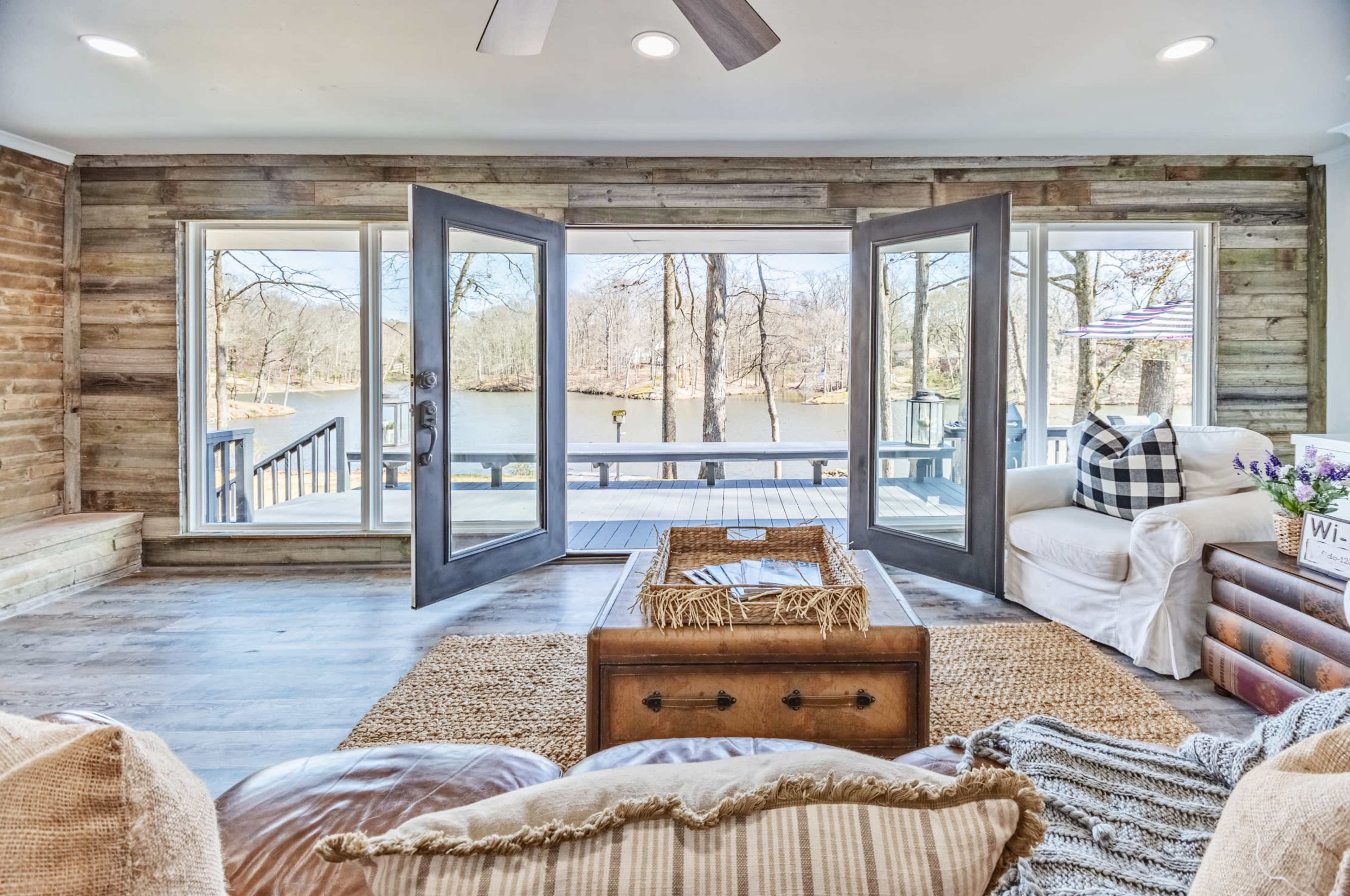 The image shows a cozy living room with large open doors leading to a deck overlooking a lake.