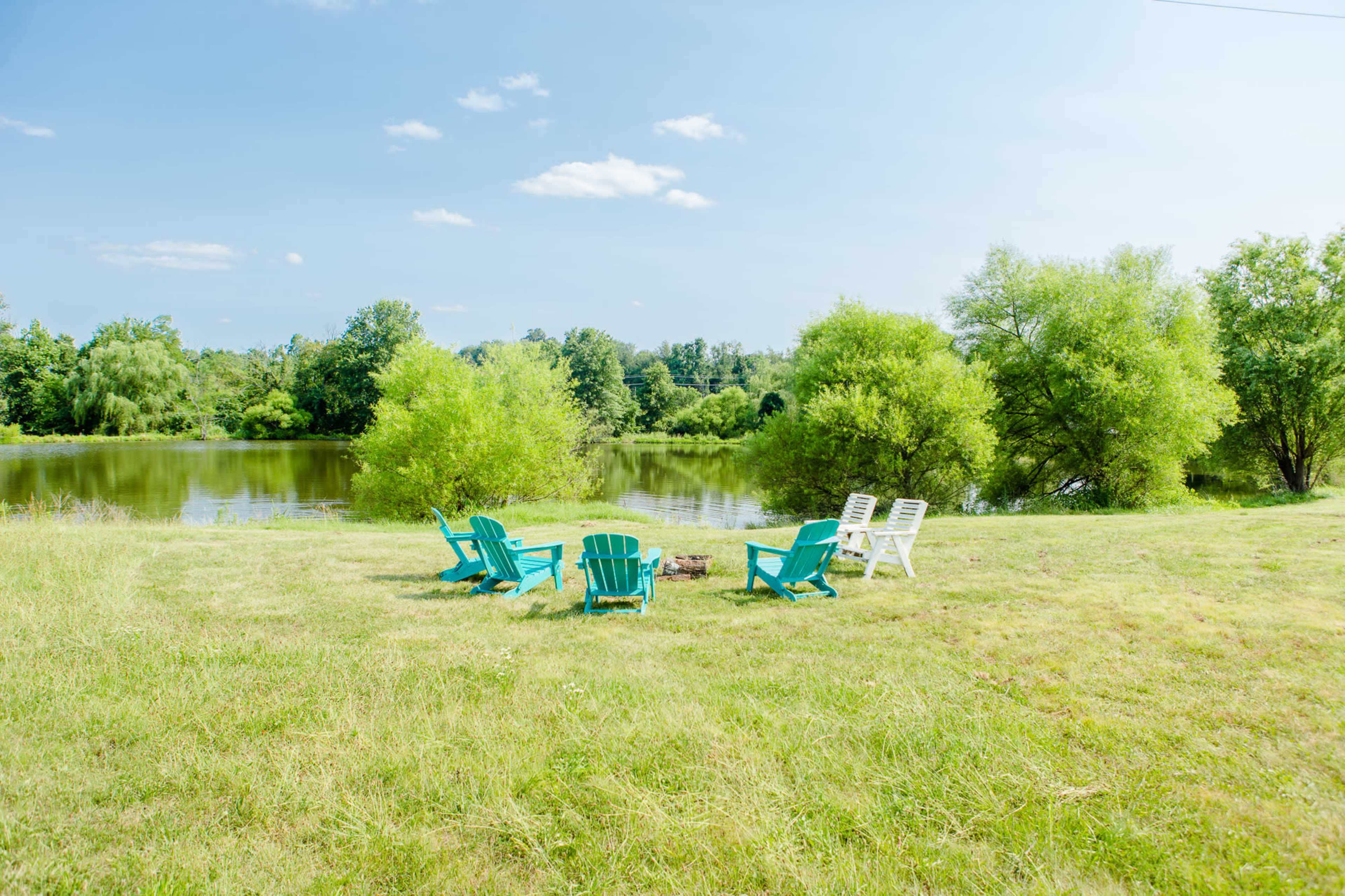 Four turquoise chairs and one white chair surround a small fire pit near a calm pond, with lush greenery in the background.