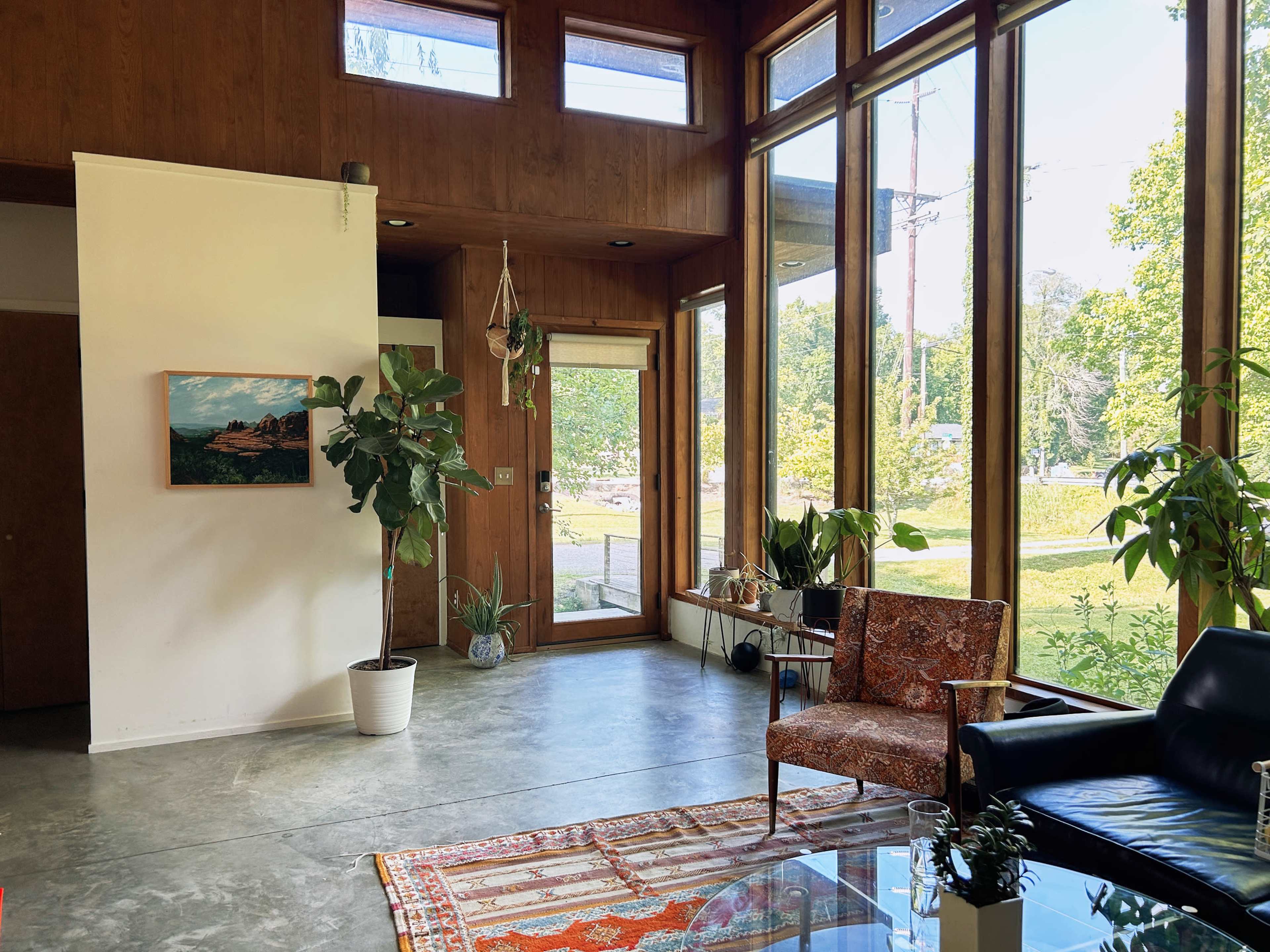 A modern living room with large windows, a potted plant, an orange and cream patterned rug, and a blend of furniture including an armchair and a glass coffee table.