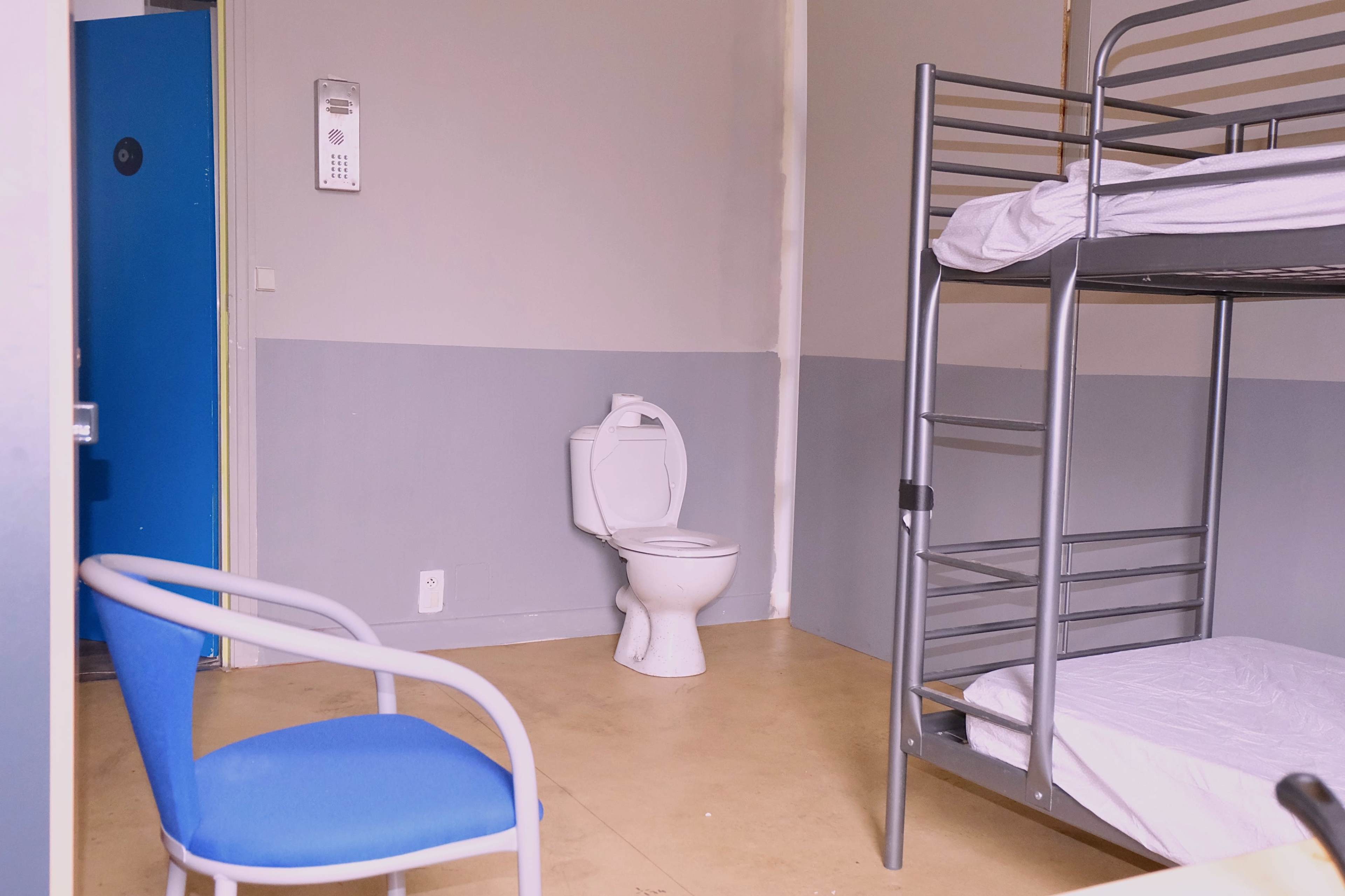 A sparse room with a blue door, a metal bunk bed with unmade mattresses, a toilet, and a blue chair.