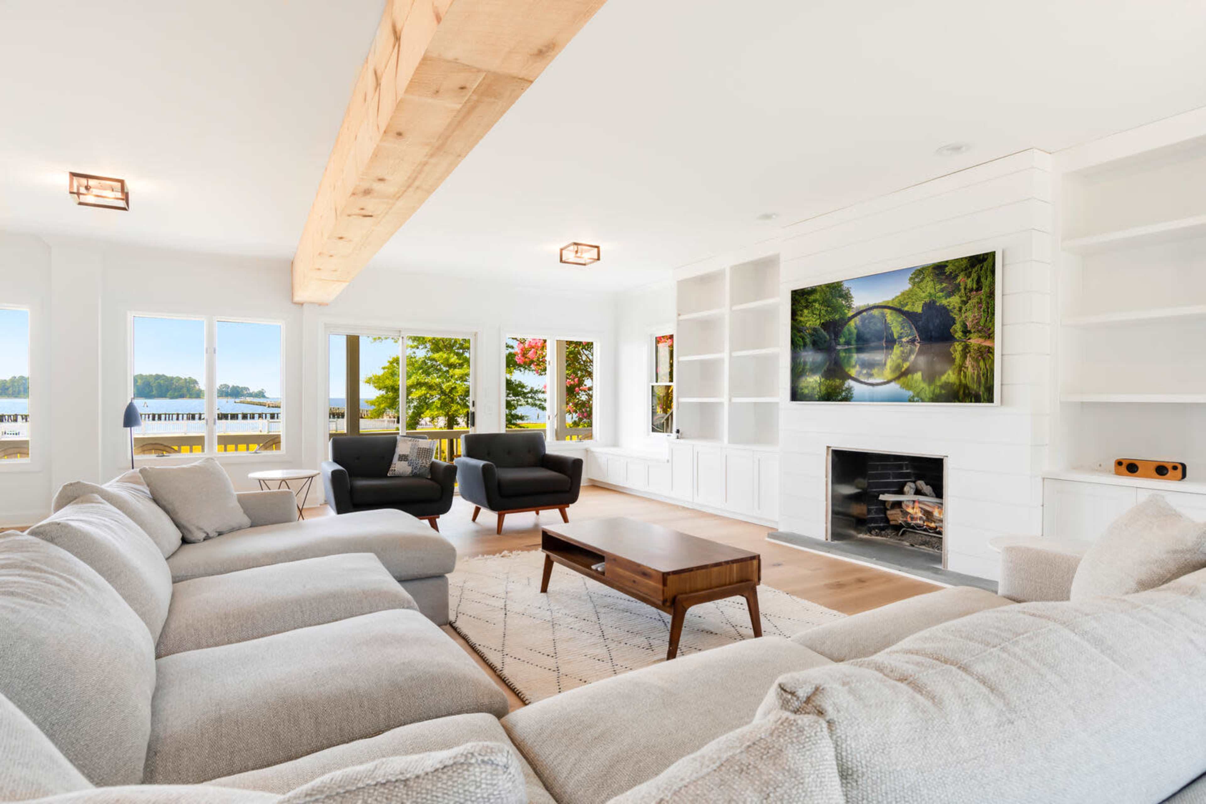 A bright, modern living room features a large sectional sofa, a coffee table, a television mounted above a fireplace, and large windows with scenic views.
