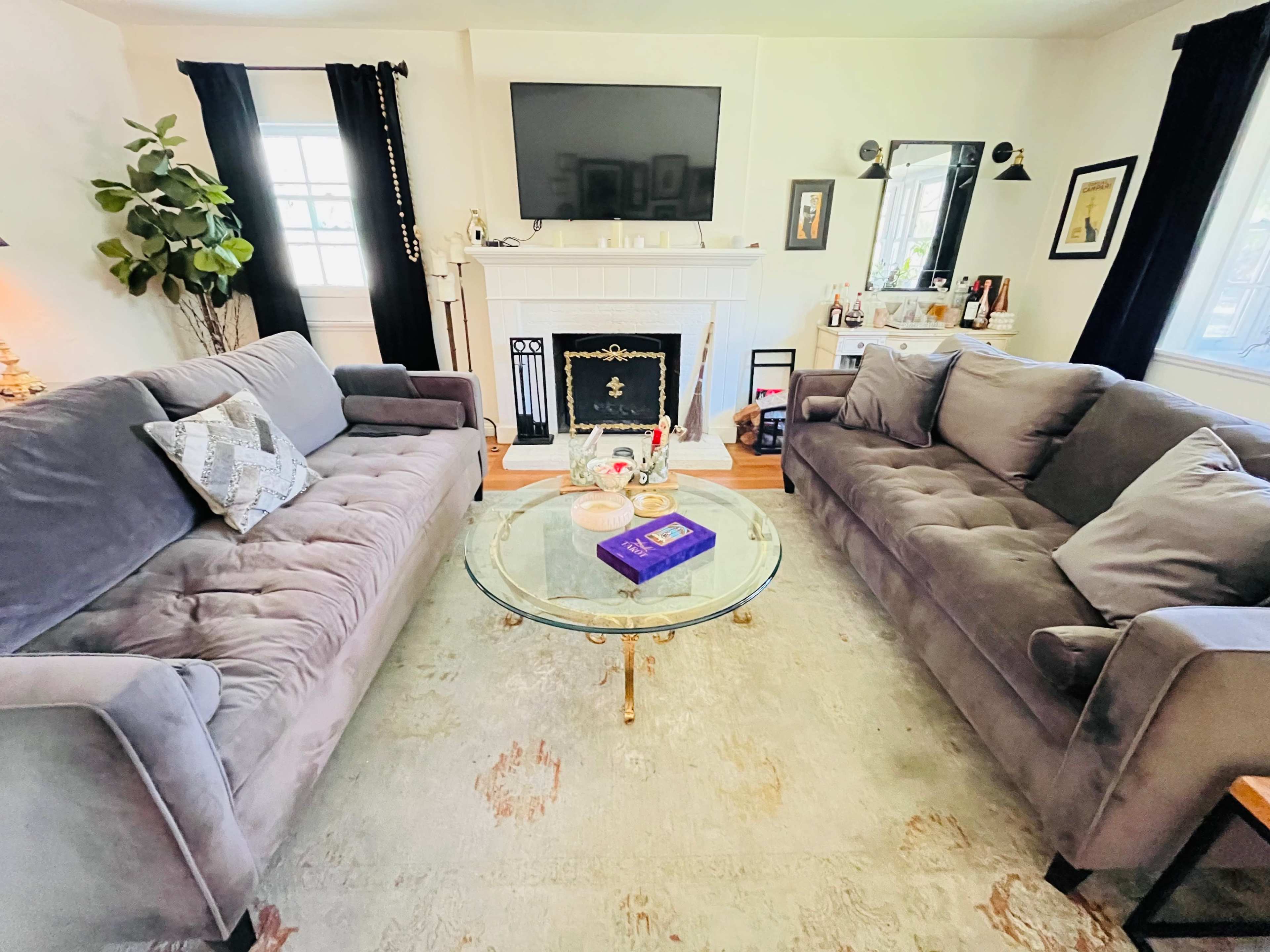 The image shows a living room with two gray sofas facing a glass coffee table and a fireplace, and a television mounted on the wall.