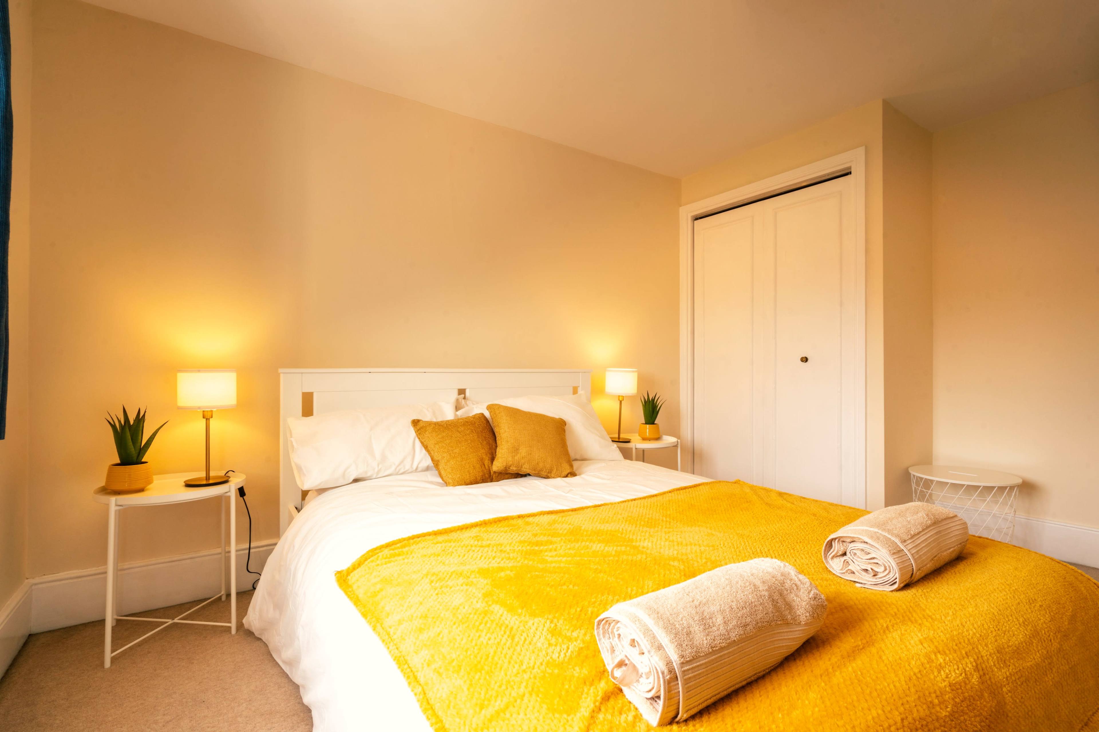 A neatly arranged bedroom with a white bed adorned with a yellow blanket, flanked by two lamps and a closet in the background.