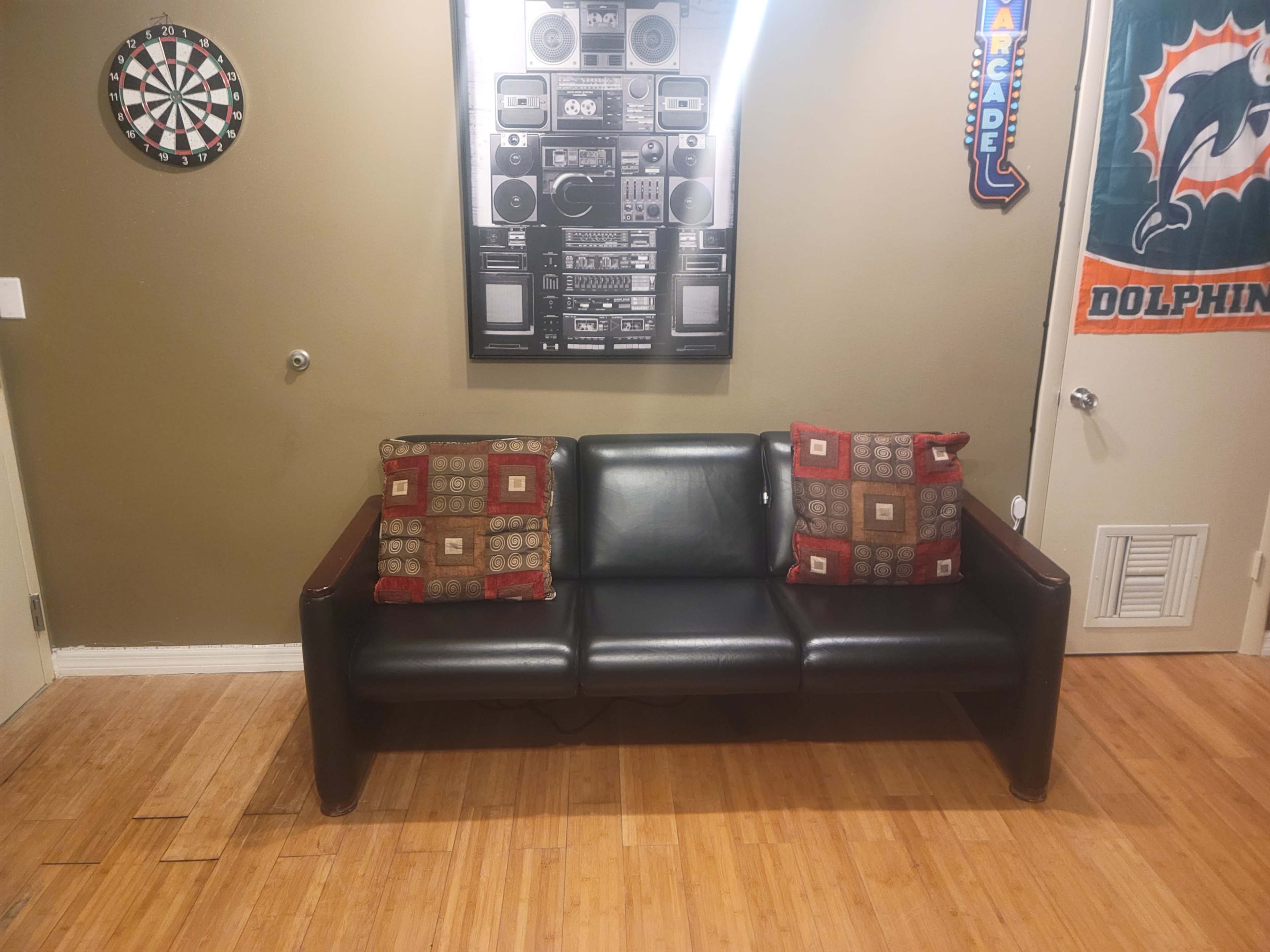 A black leather sofa with two patterned cushions against a beige wall, with a dartboard and a vintage stereo system poster hanging above it.