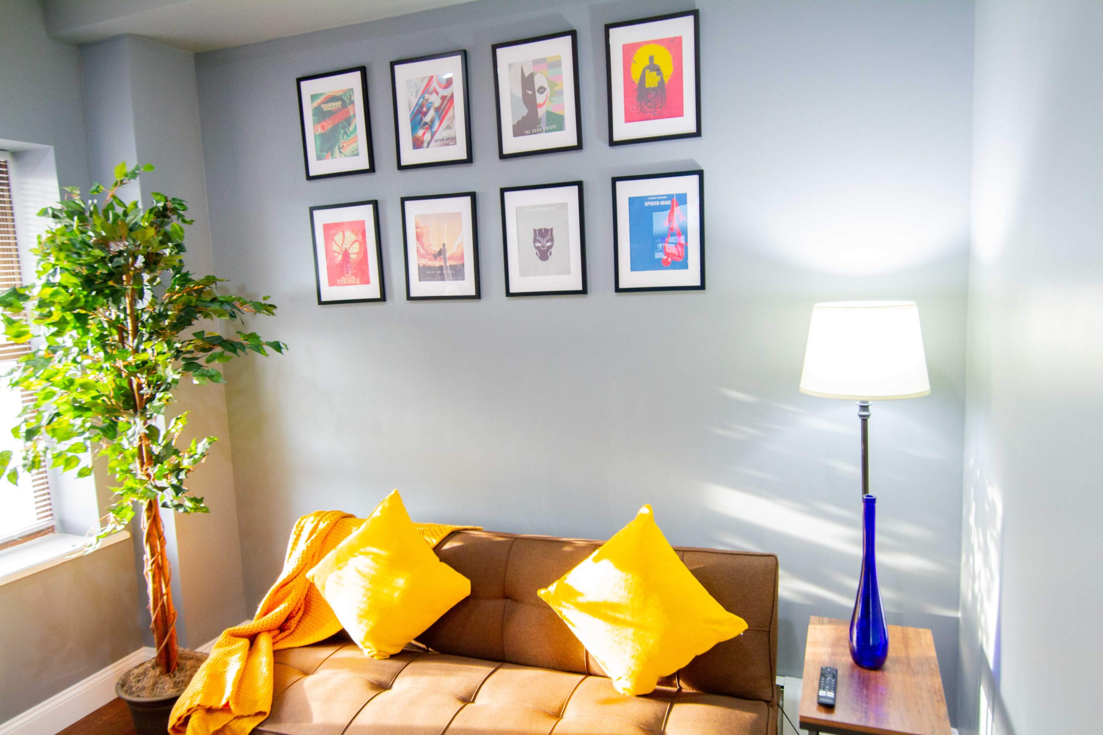 A small living area with a brown sofa adorned with yellow cushions, a blue lamp, and a wall displaying an arrangement of framed art prints.