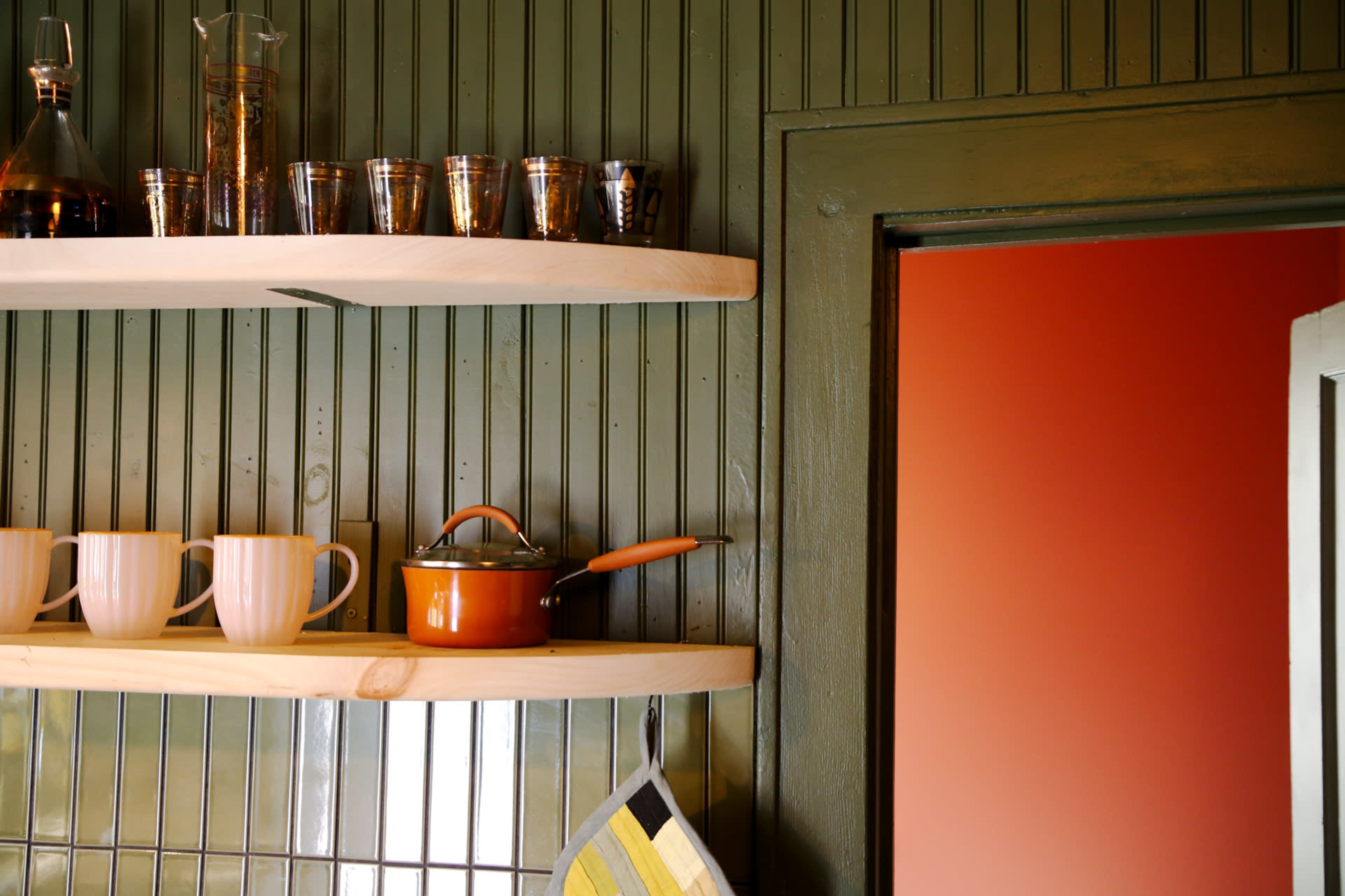 A small kitchen space features wooden shelves holding glassware and a small orange pot, with a partially open door showing a reddish wall.
