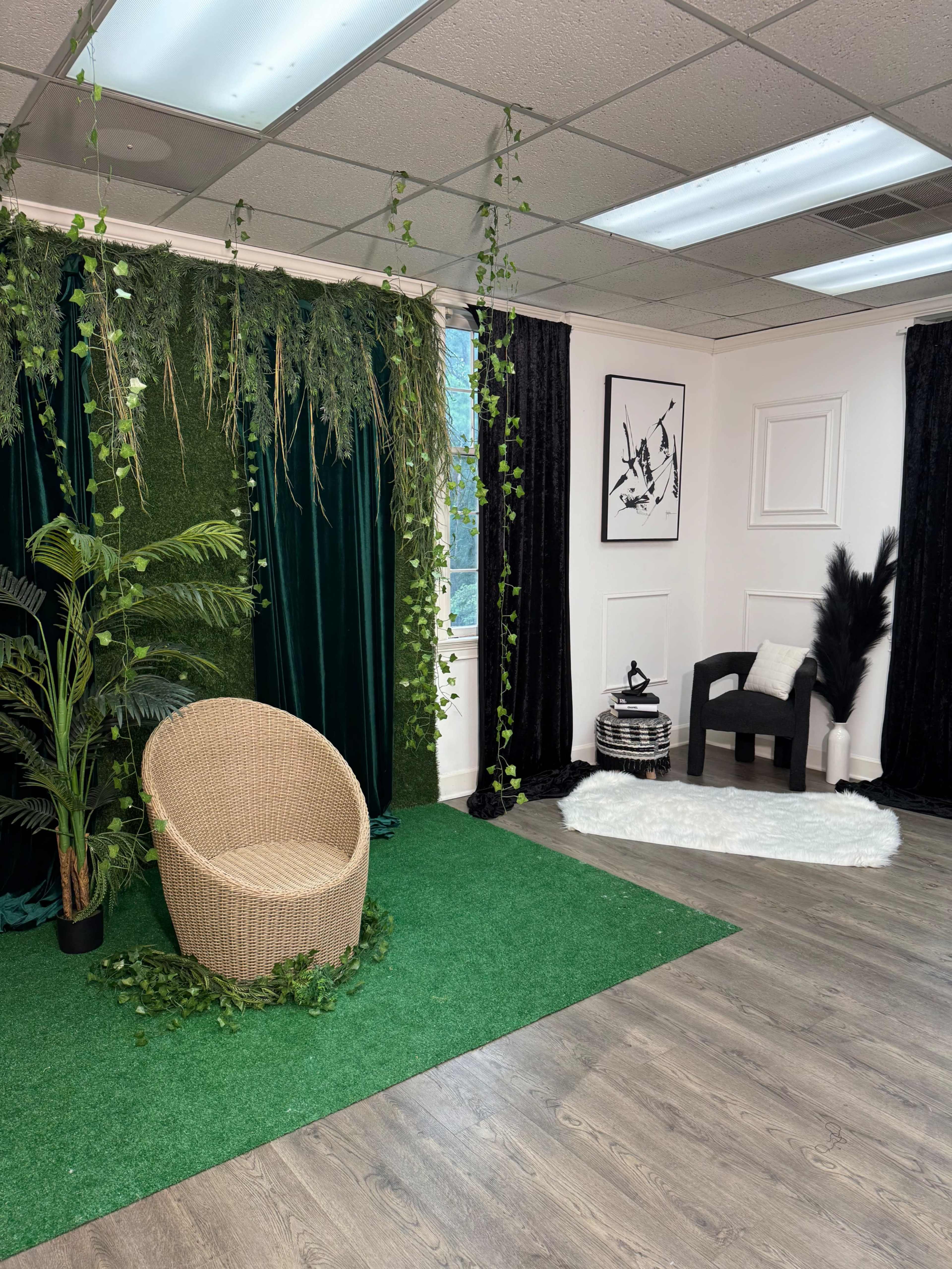 The image shows a studio space with a green carpet, a wicker chair surrounded by plants, and contrasting black curtains forming a backdrop.