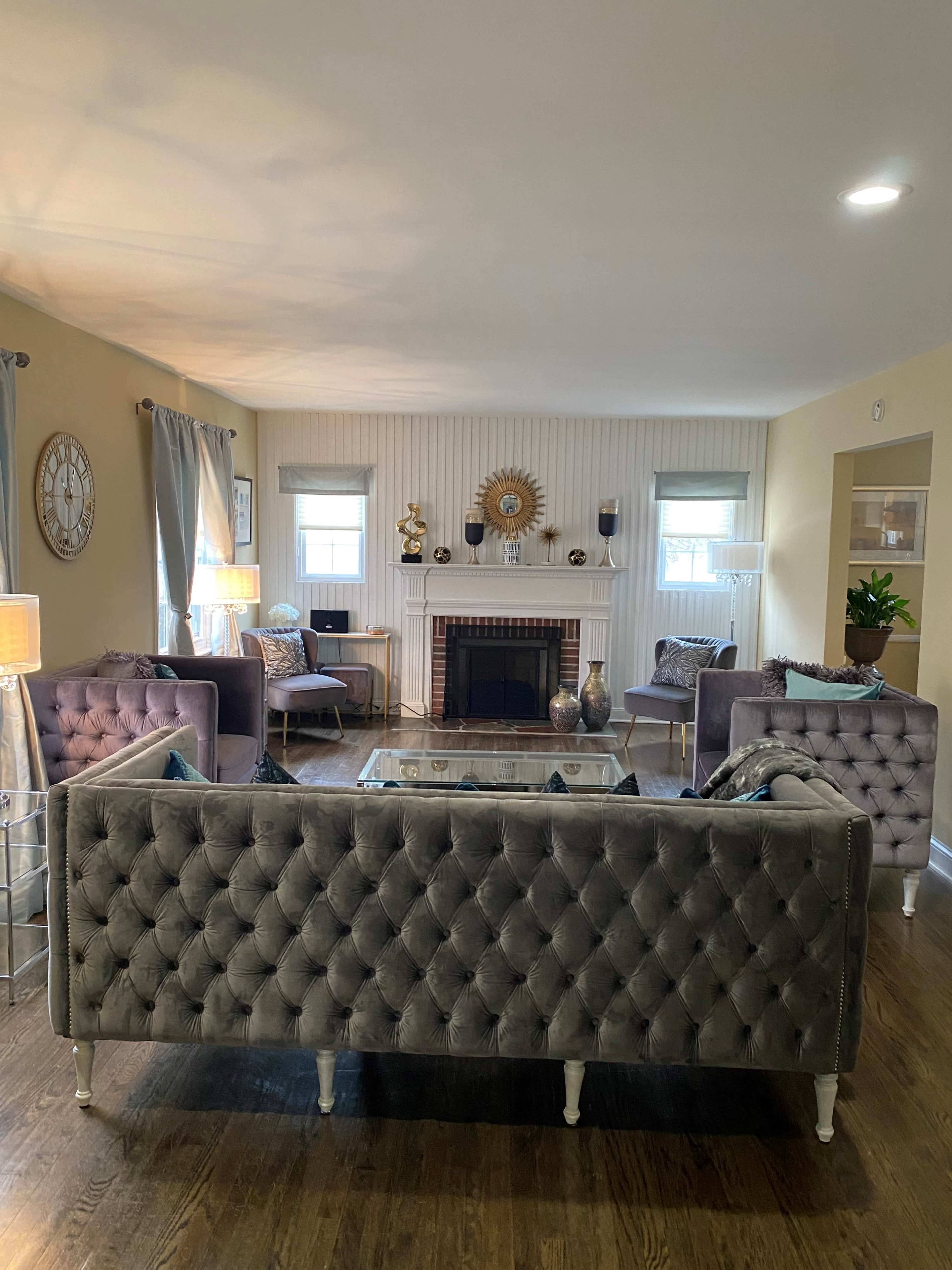 A living room features tufted gray sofas, a glass coffee table, a decorative fireplace, and large windows with light-colored curtains.