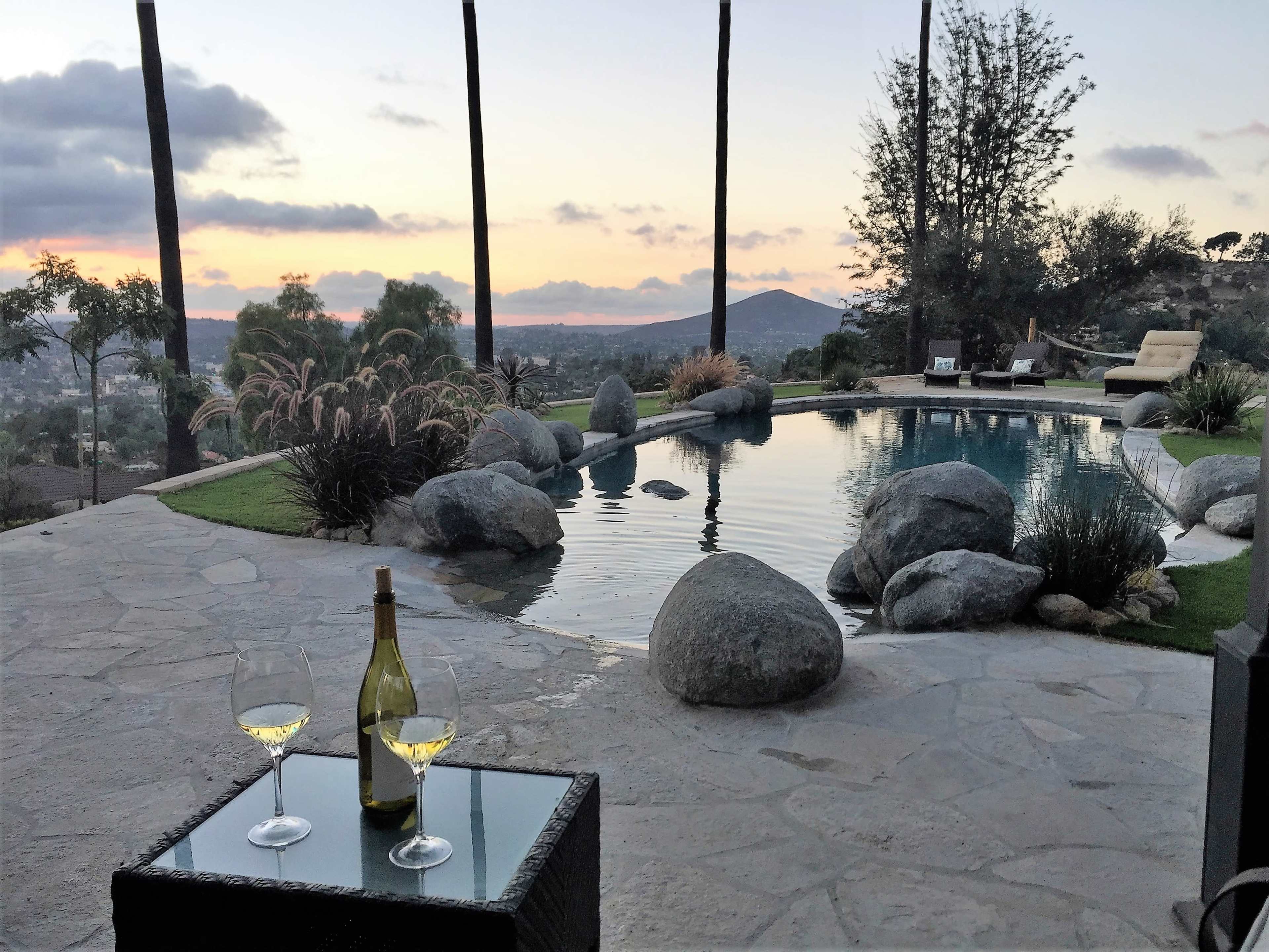 The image shows a serene poolside scene at sunset, featuring a glass table with two wine glasses and a bottle, surrounded by rocks and greenery.