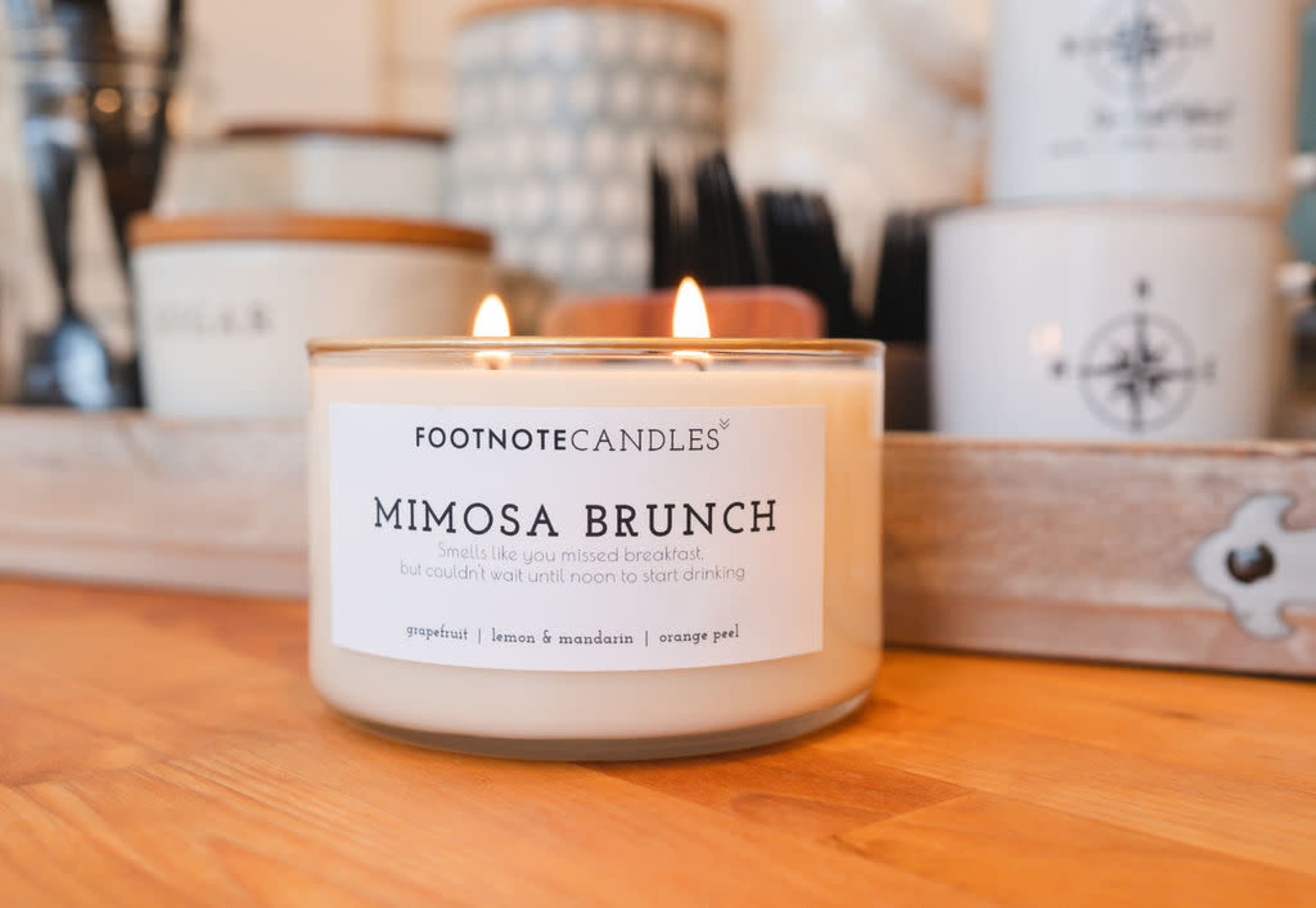 A lit candle labeled "Mimosa Brunch" from Footnote Candles sits on a wooden tray surrounded by various small containers.