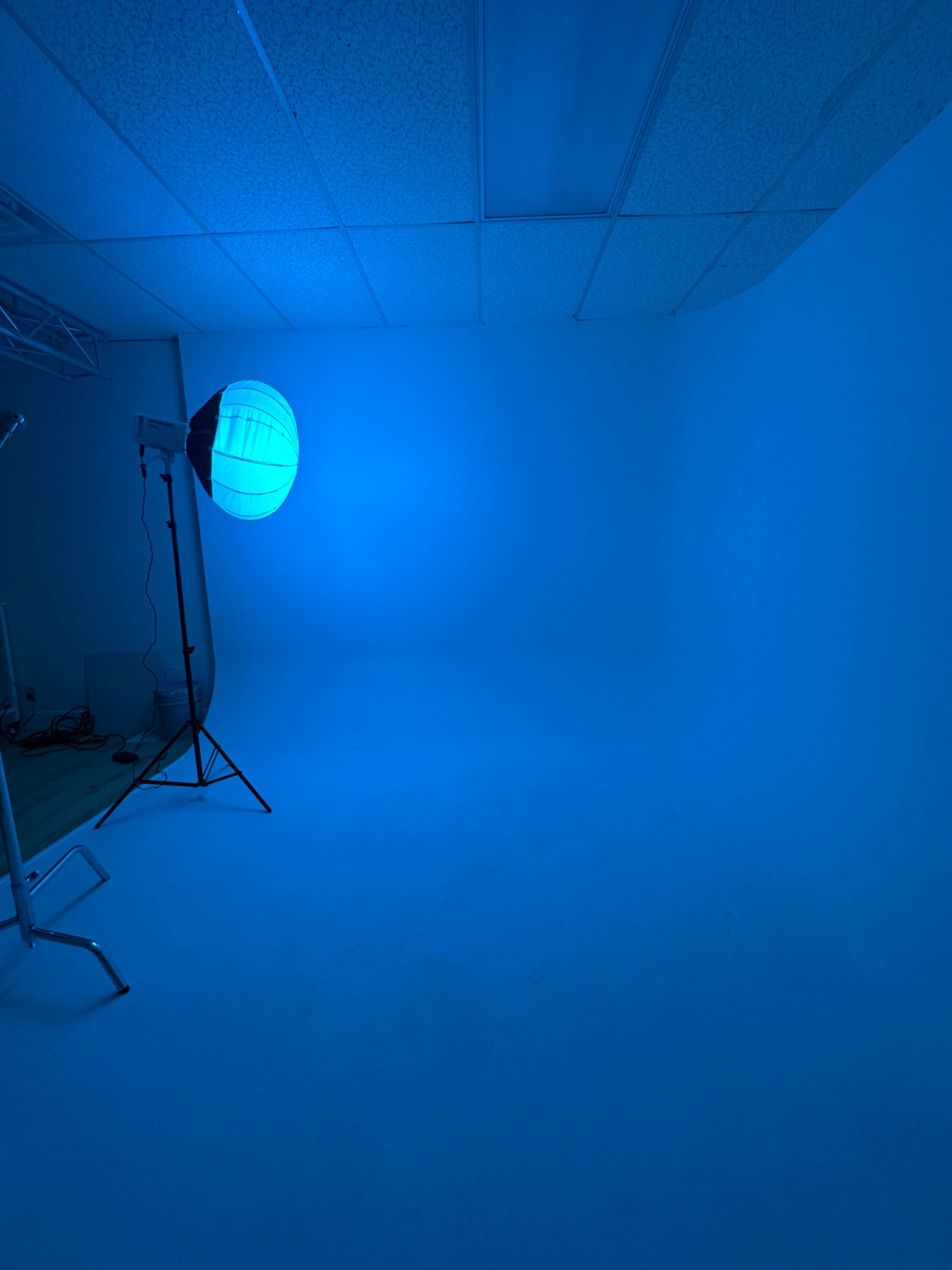 The image shows a photography studio with a blue backdrop and a strobe light on a stand.