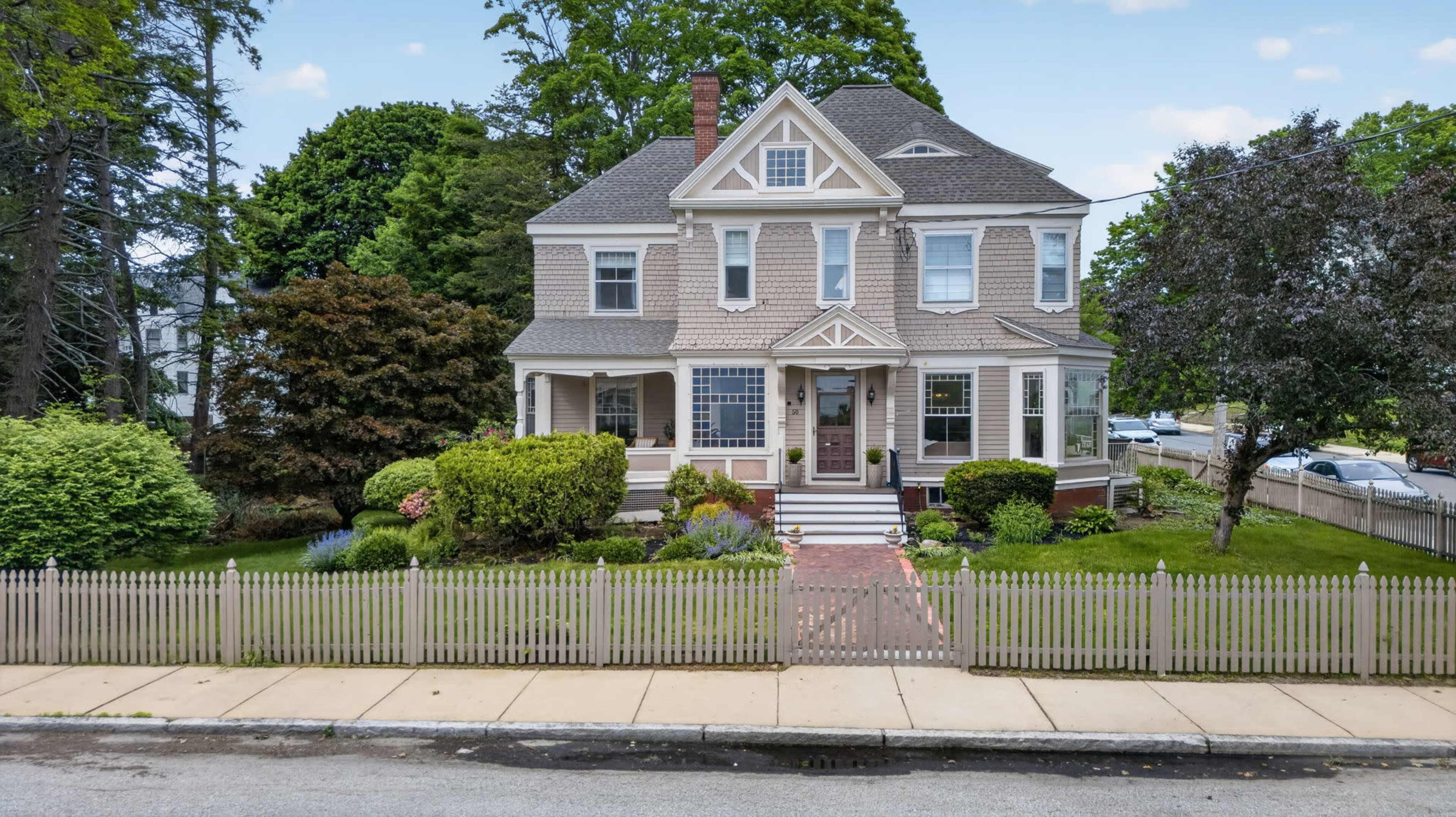 A large, historic house with a front porch and a well-kept yard is surrounded by a picket fence.