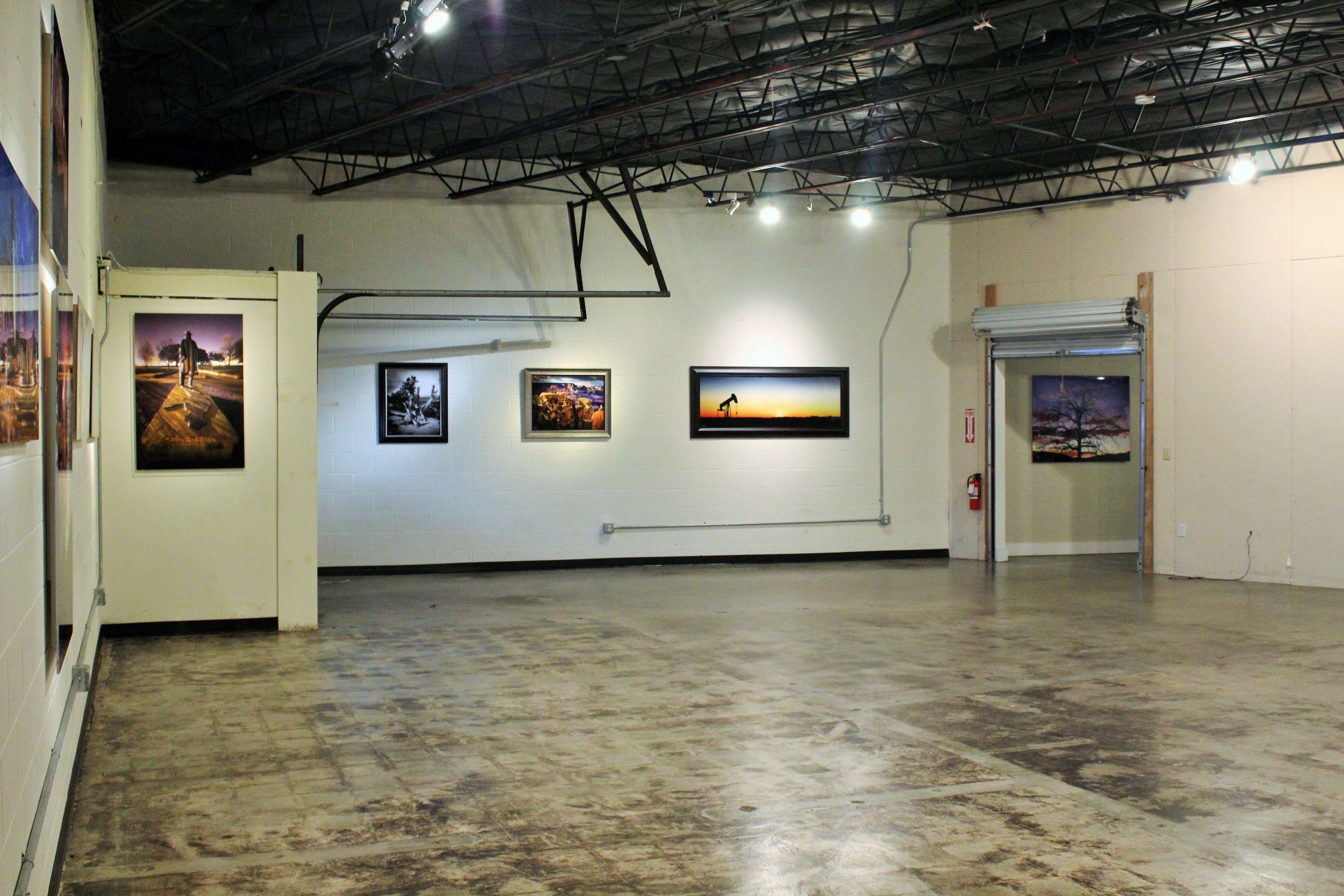 An empty gallery space with several photographs displayed on the walls and a metal roll-up door on one side.