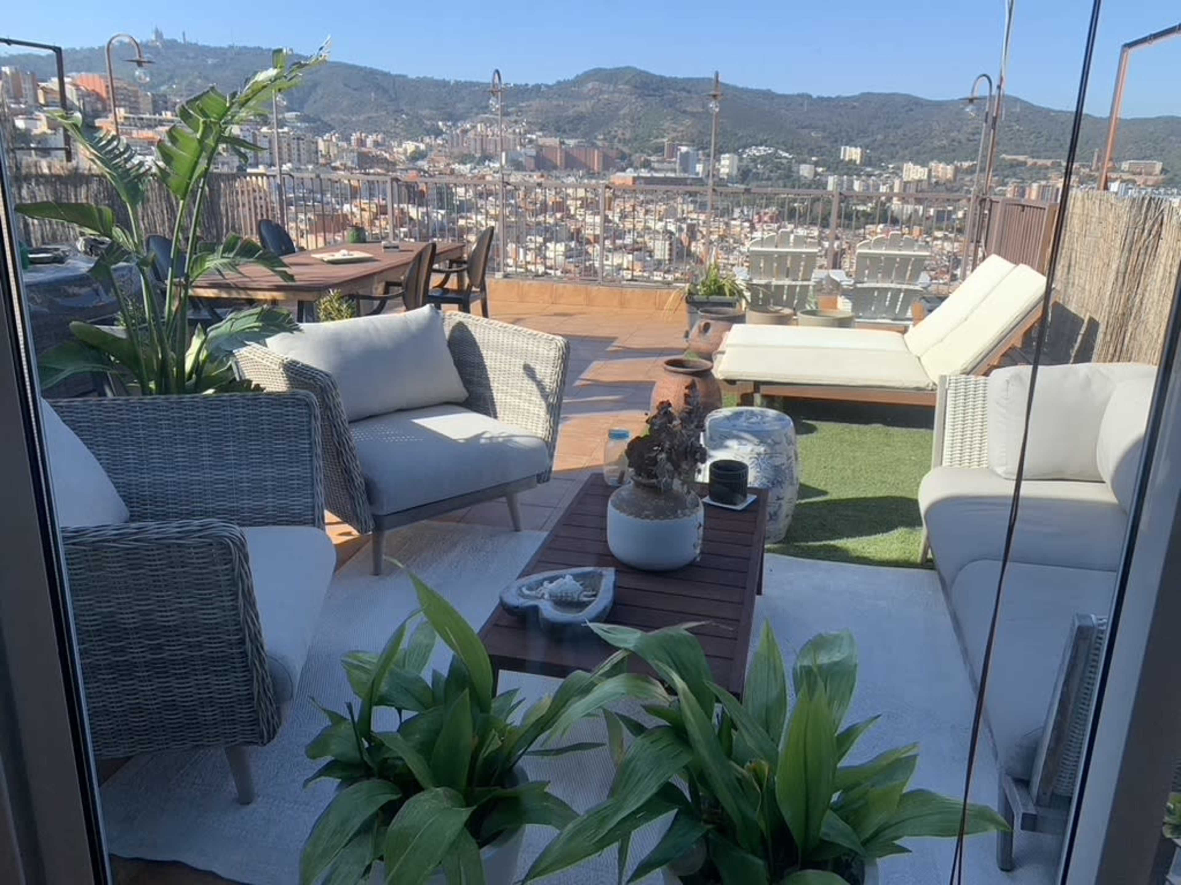 A rooftop terrace with seating areas, potted plants, and a view of a city landscape in the background.