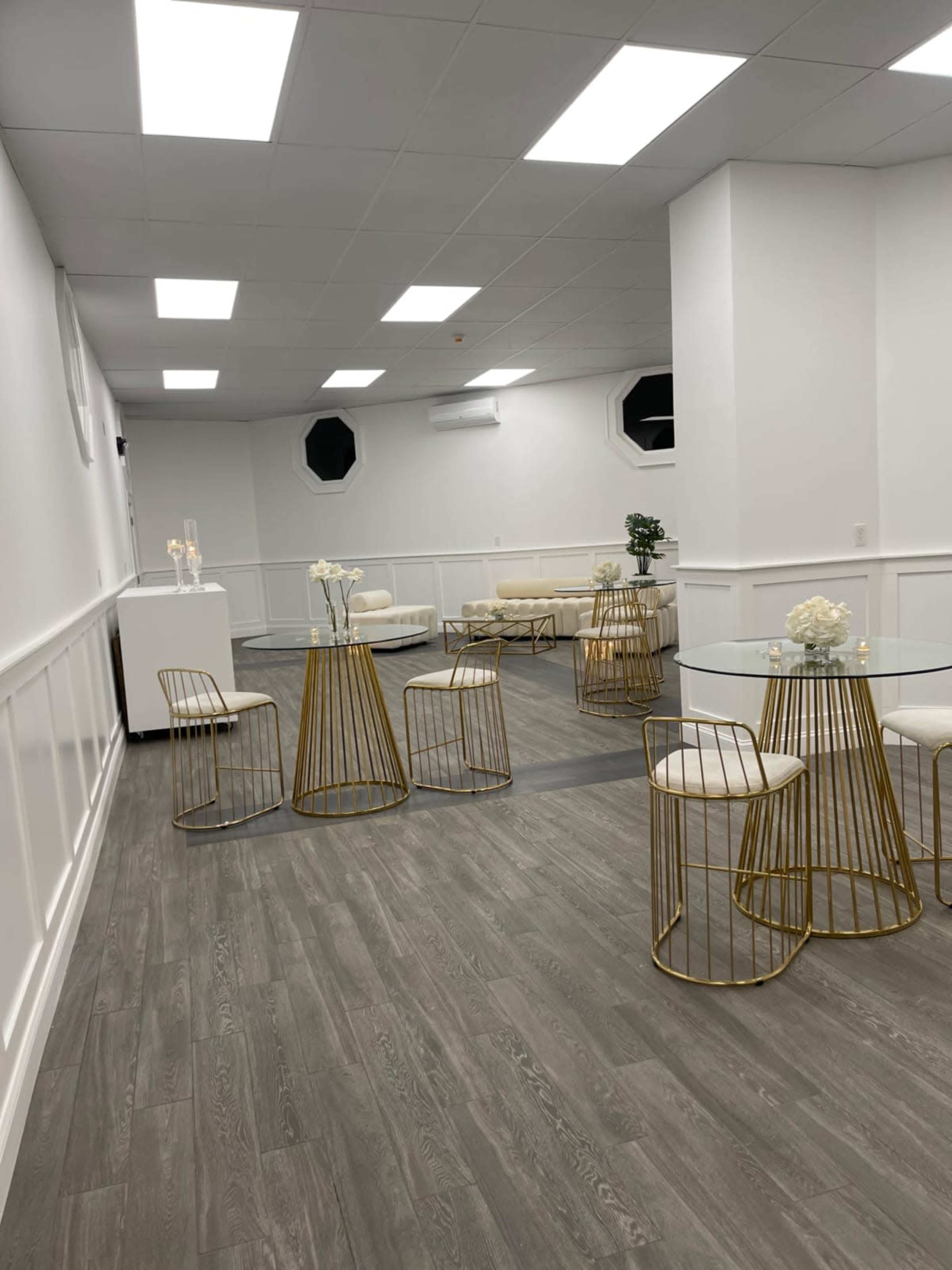 A modern, minimalist event space with white walls, a wooden floor, and gold-accented tables and chairs arranged around the room.