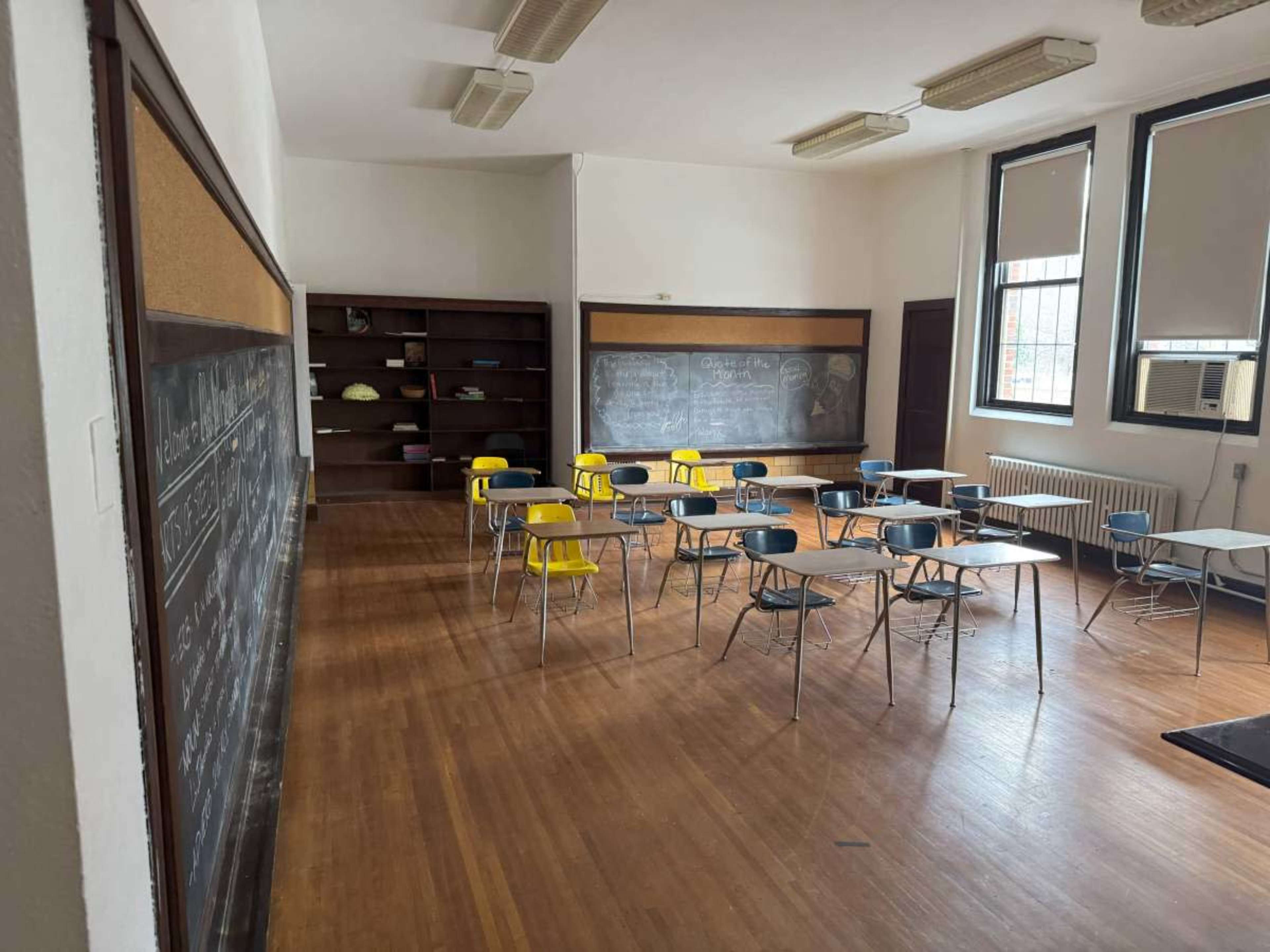 The image shows an empty classroom with several desks arranged in rows and chalkboards on the walls.