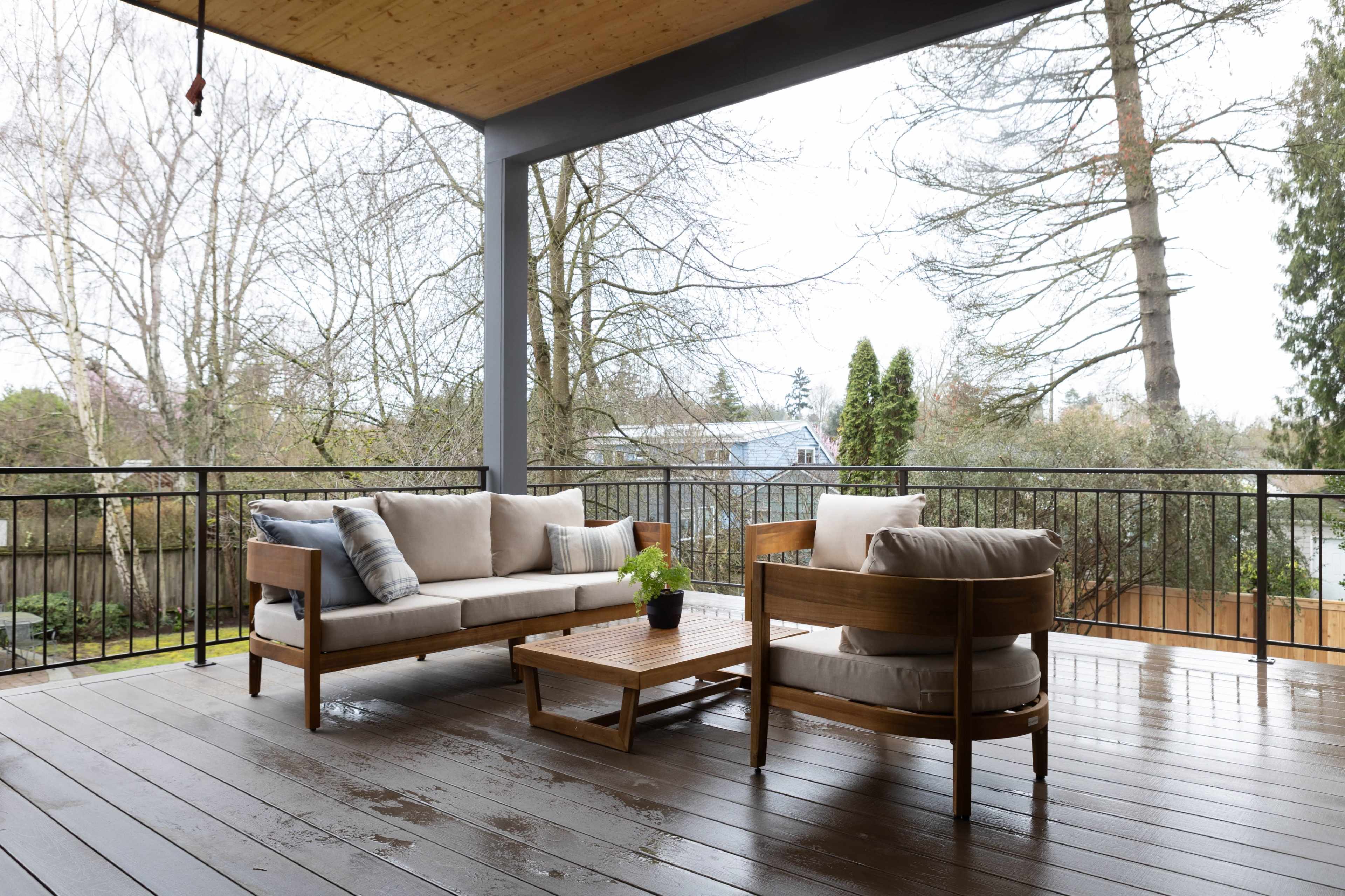 A covered outdoor patio features a wooden furniture set with cushions, a coffee table, and a lush green backdrop of trees.