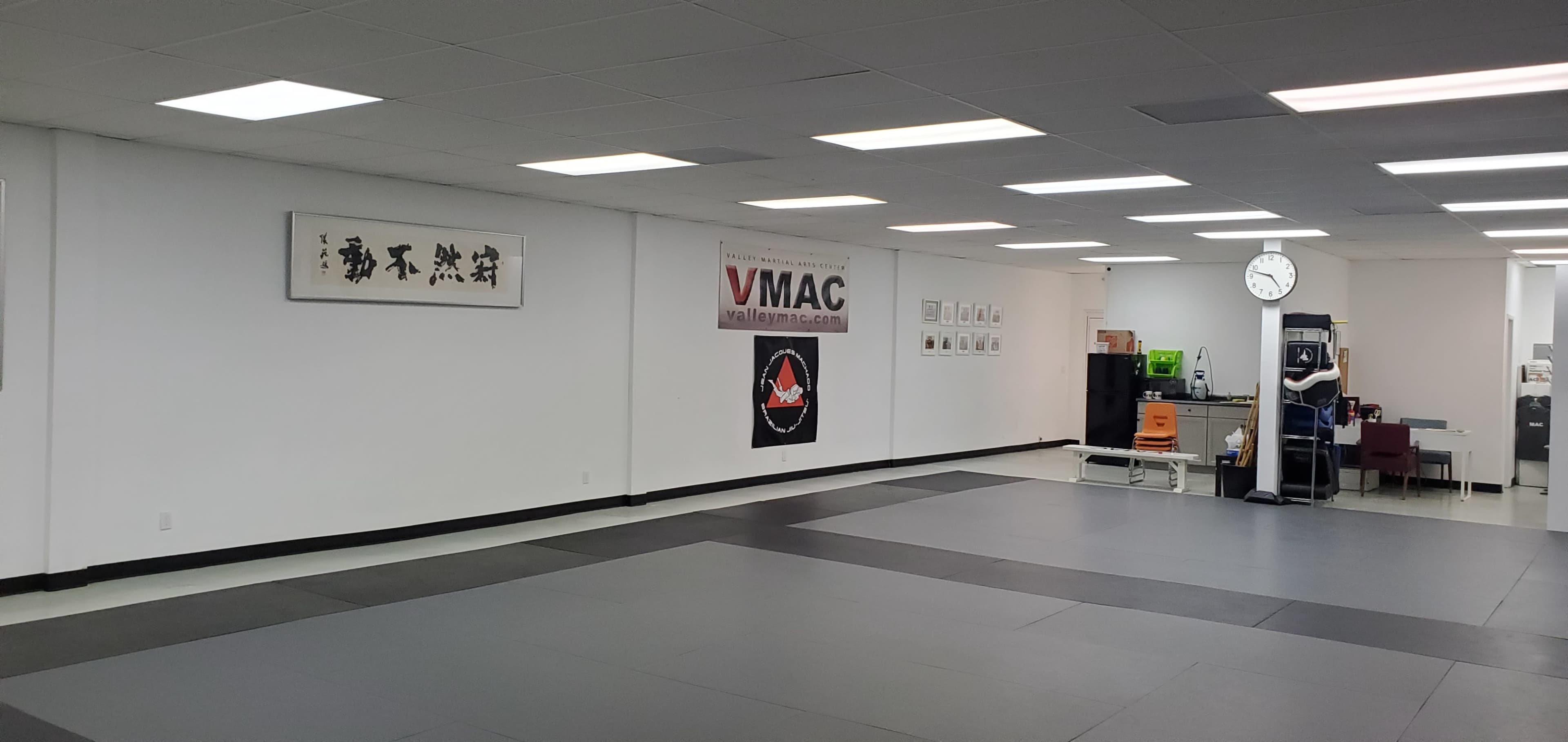 A martial arts training studio with a large open floor covered in mats, white walls, and various wall decorations, including a clock and a promotional banner.