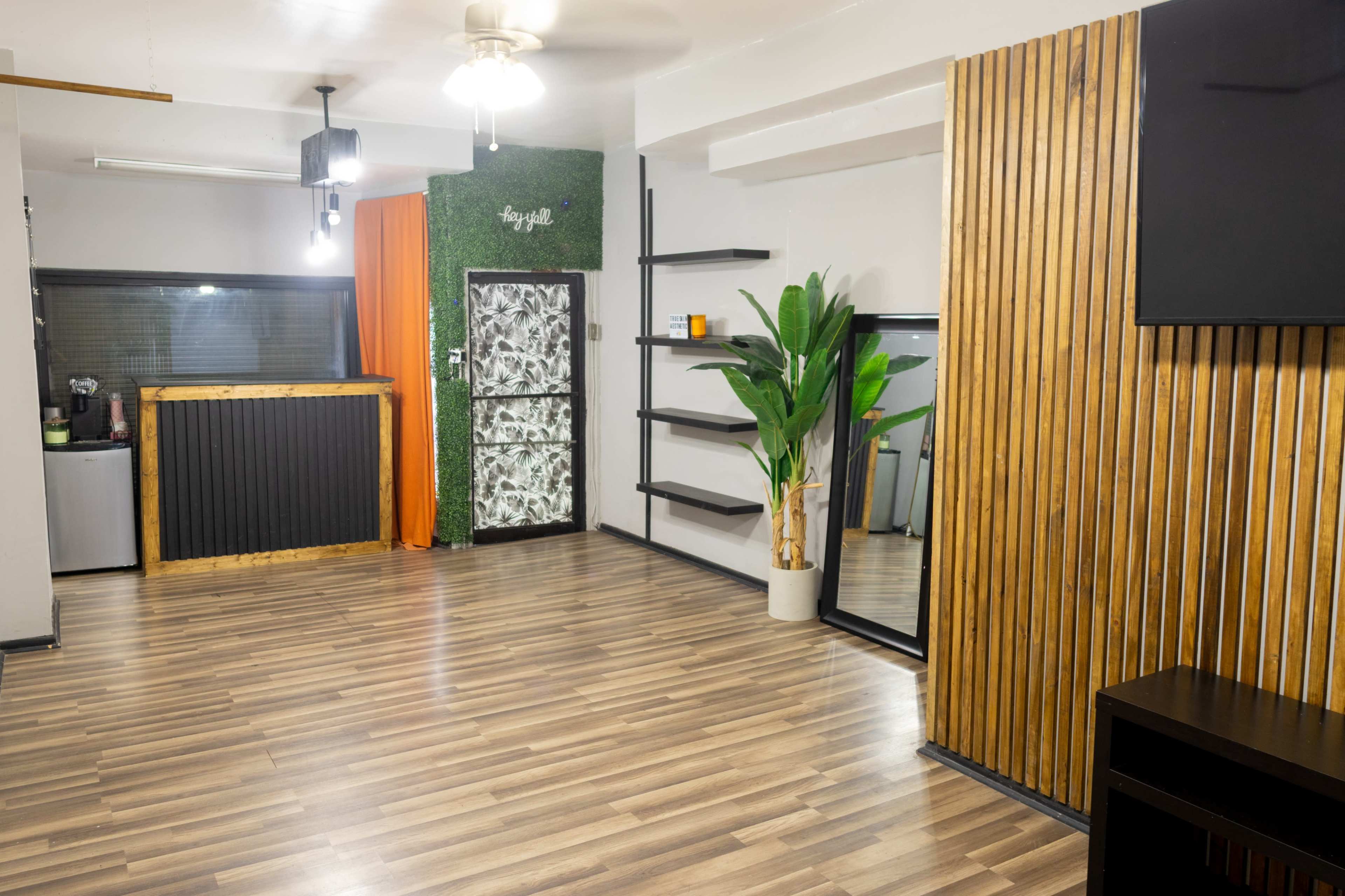 The image shows a spacious, modern room with wooden flooring, featuring a green accent wall, a large mirror, and several shelves next to a plant.