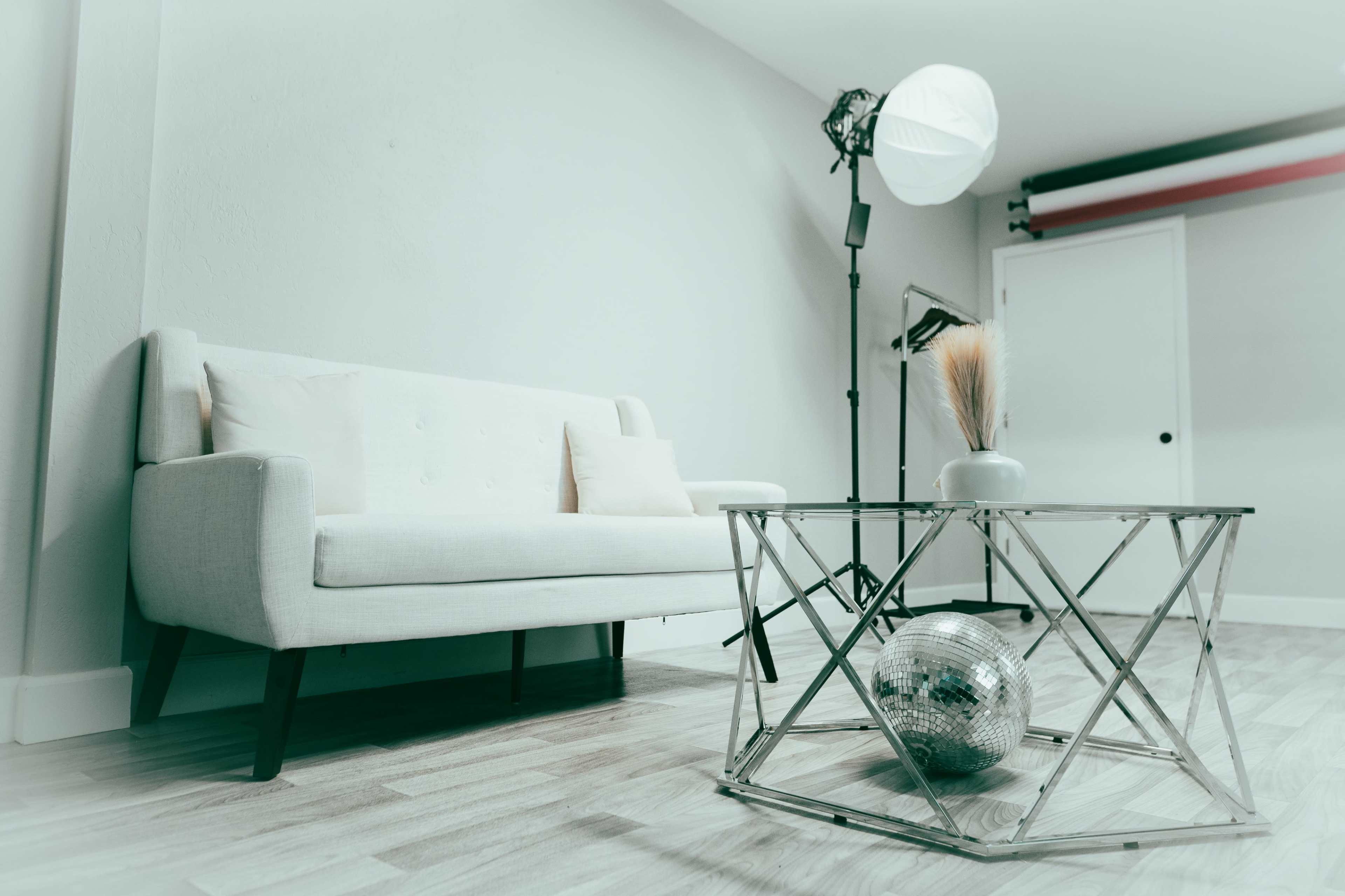 A light-colored sofa is positioned in a room with a reflective glass table that holds a decorative object, while a softbox light stands nearby.