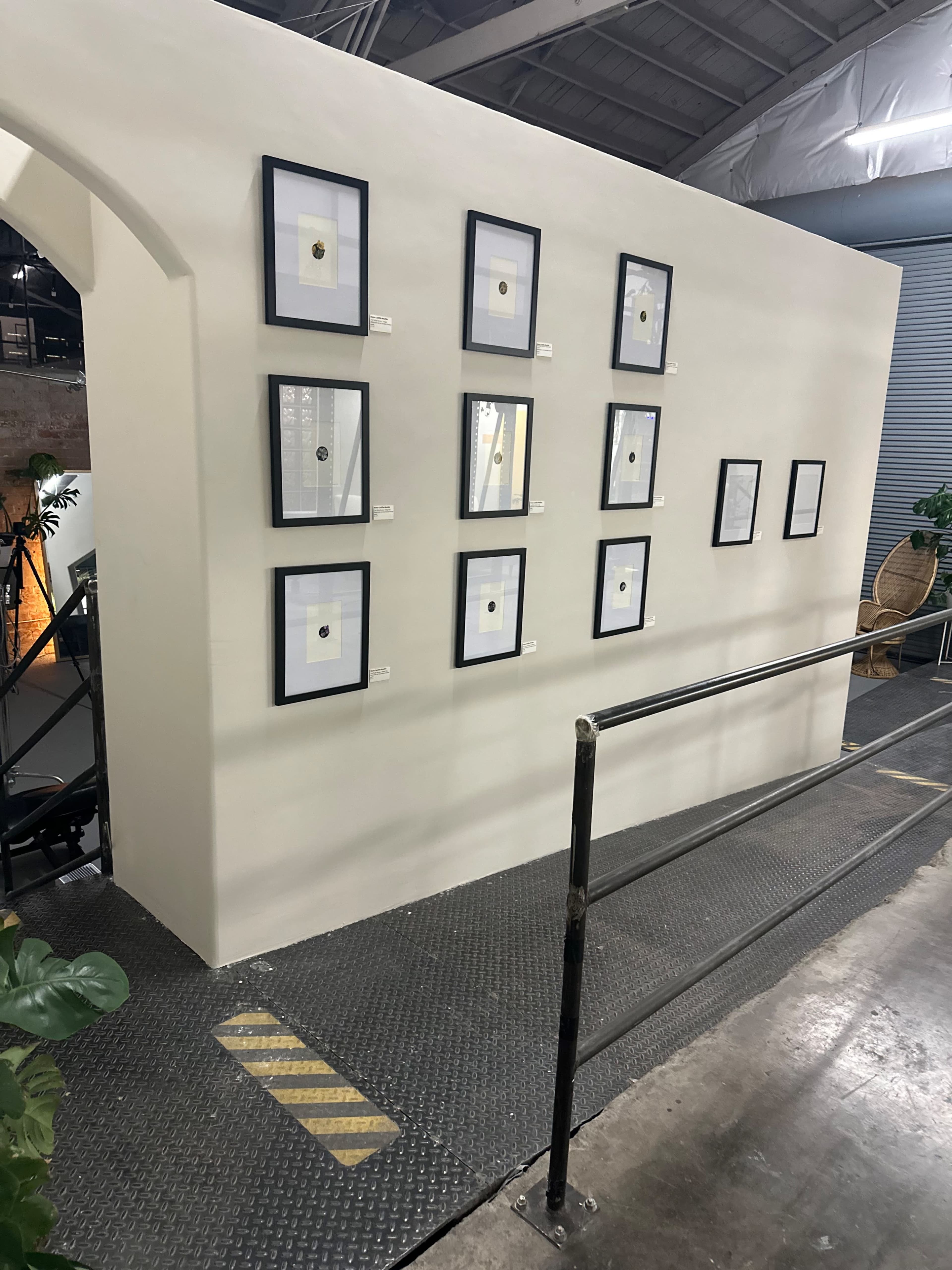 A gallery wall with nine framed artworks arranged in a grid on a white partition in a spacious, industrial setting.