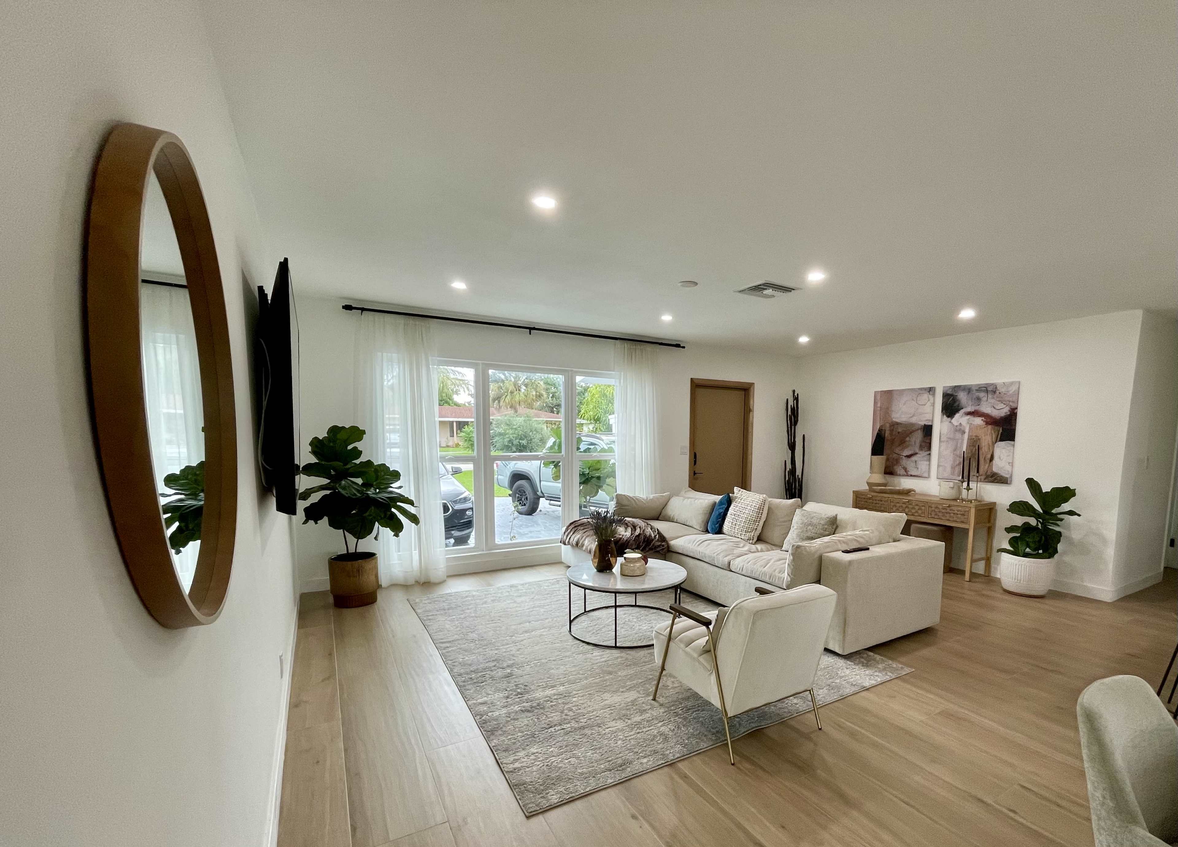 The living room features a light-colored sectional sofa, a coffee table, and large windows with sheer curtains, allowing natural light to illuminate the space.