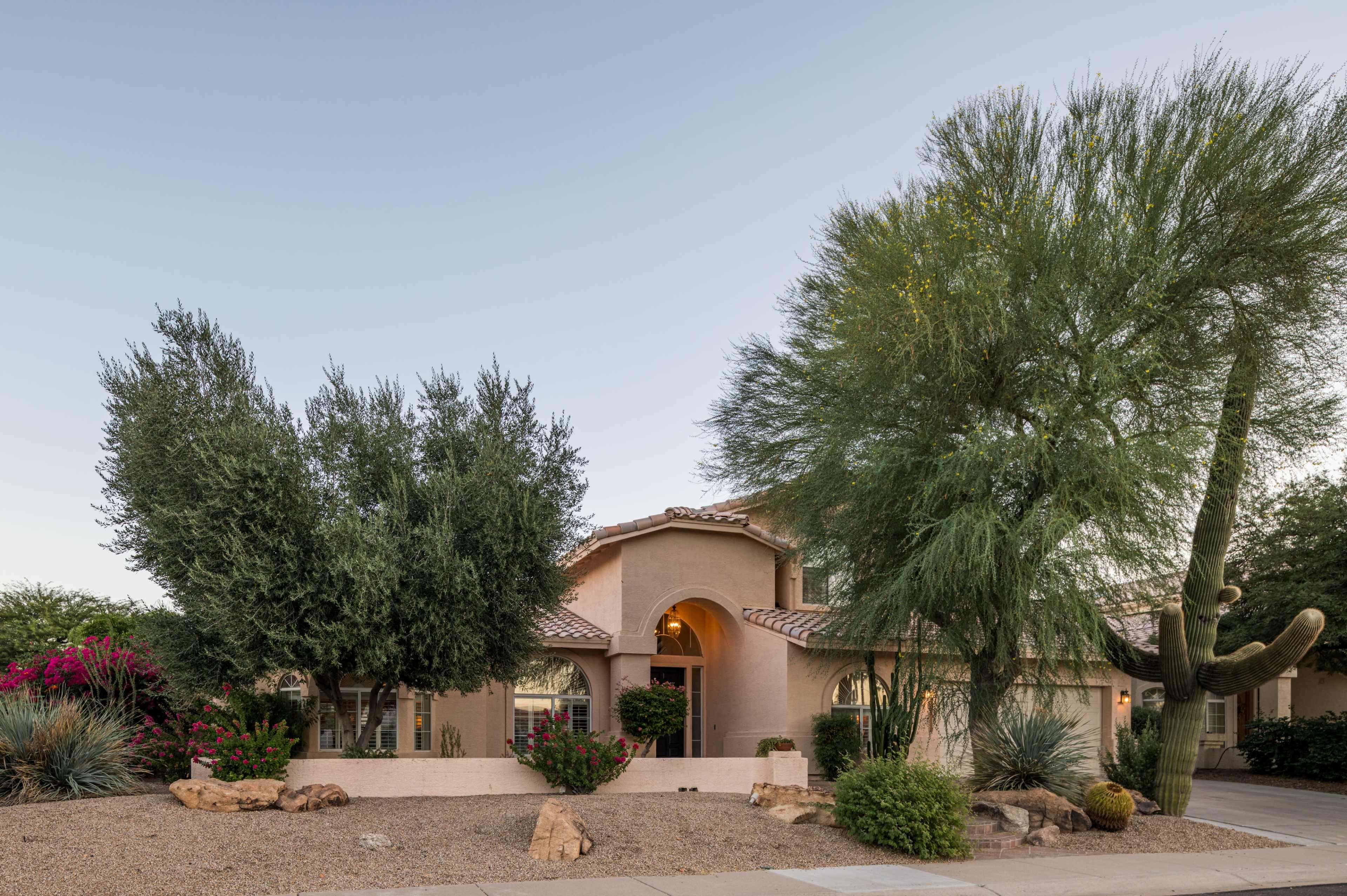 A two-story desert home with a tile roof, surrounded by various shrubs and trees, set against a clear sky.