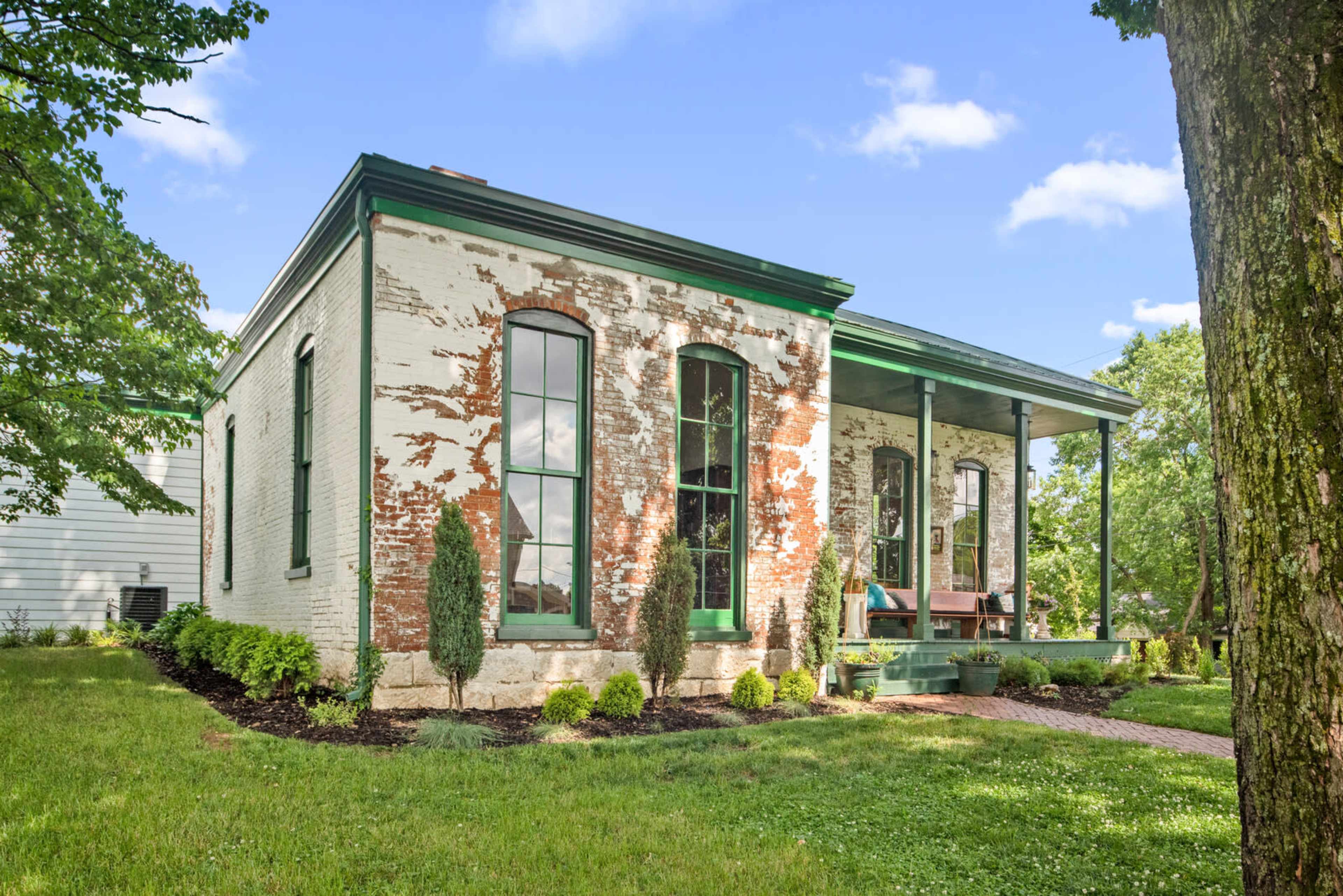 A historic brick house with large windows and a green porch surrounded by a grassy yard and trees.
