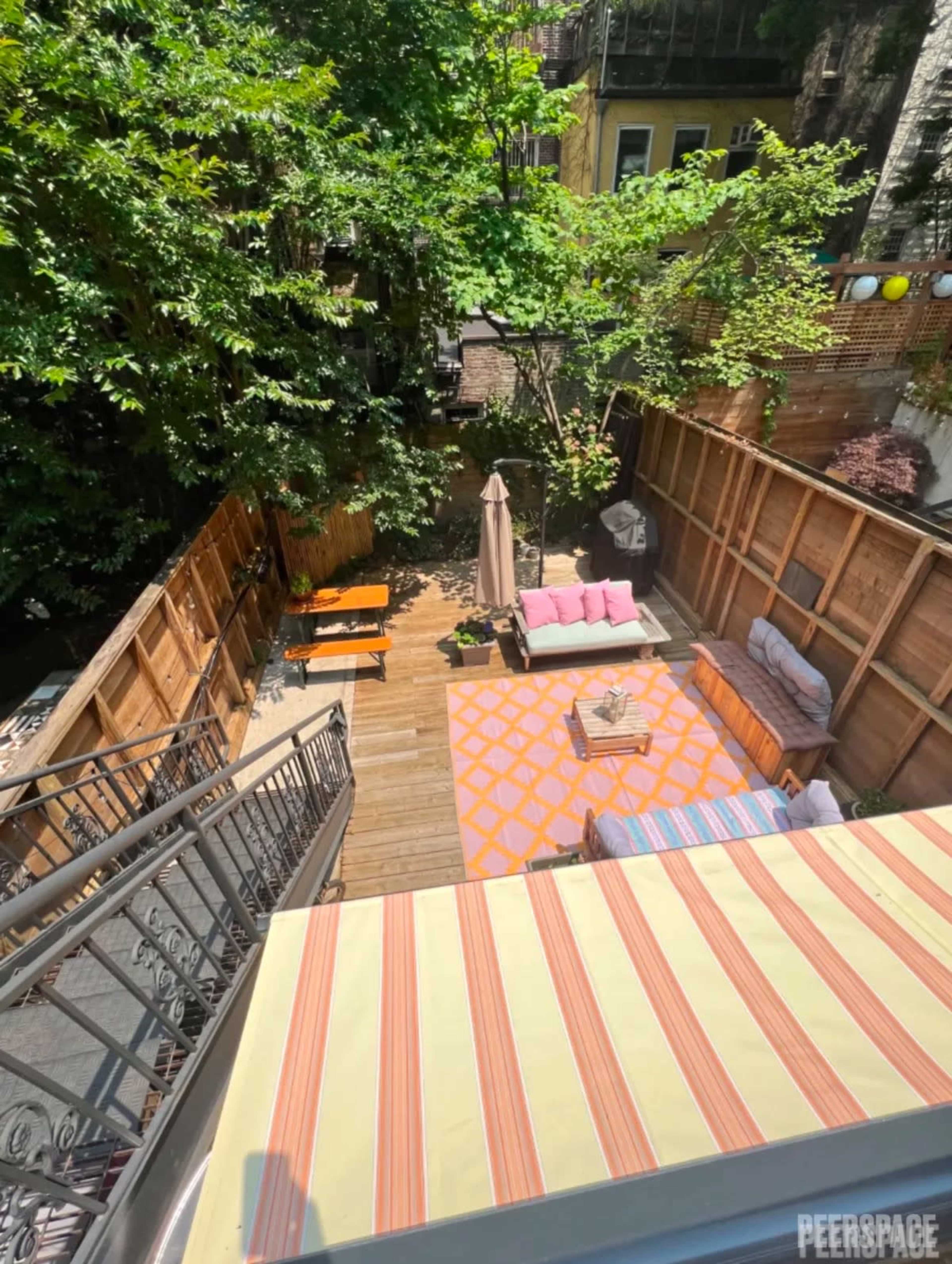 The image shows a landscaped outdoor patio area with wooden fencing, seating arrangements, a table, and a large umbrella, surrounded by trees.