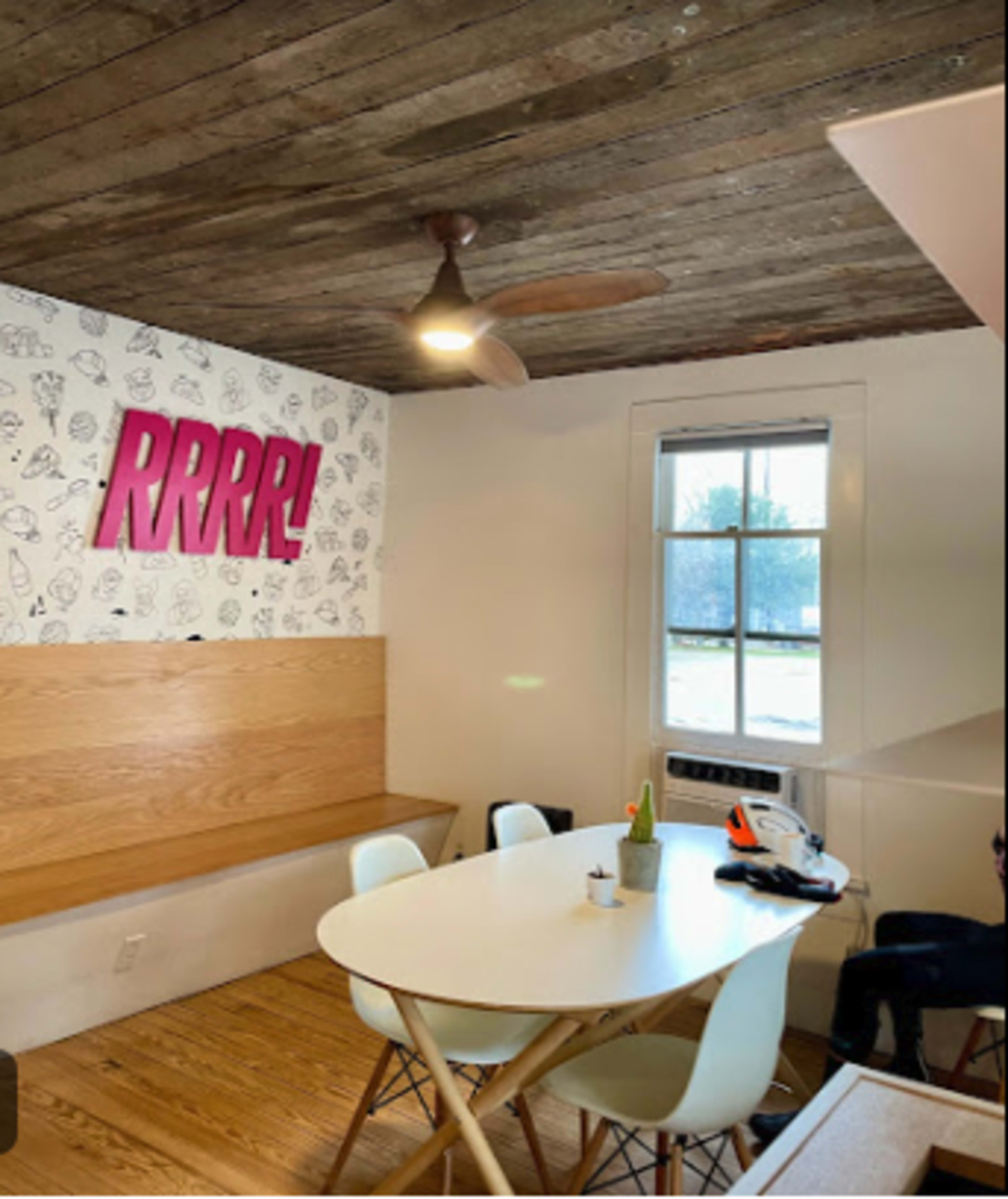 A dining area features a wooden ceiling, a white table surrounded by chairs, and a wall with pink decor and a window.