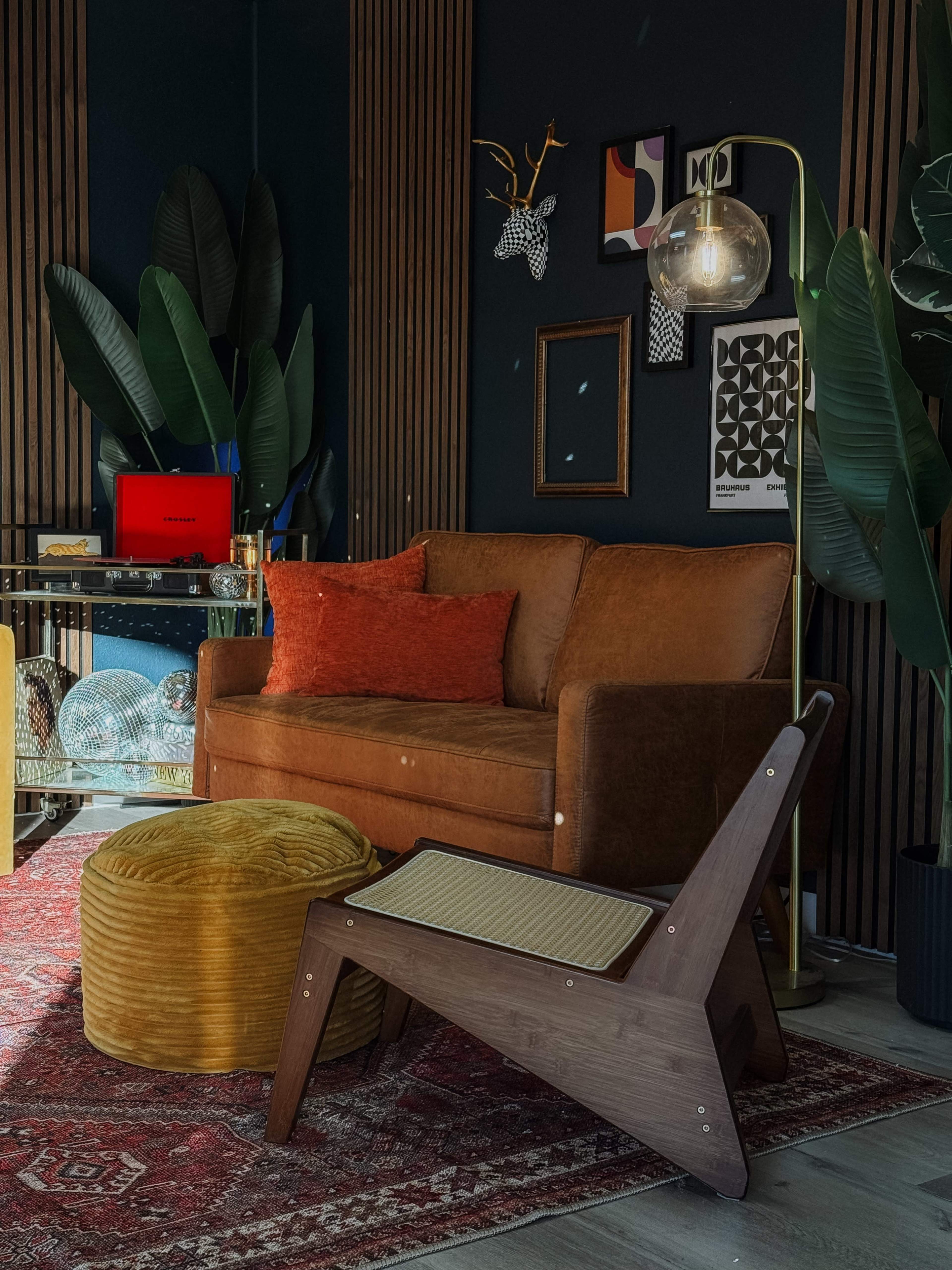The image shows a cozy living room corner featuring a brown sofa, an orange cushion, a yellow pouf, a wooden chair, and decorative plants against a dark wall.