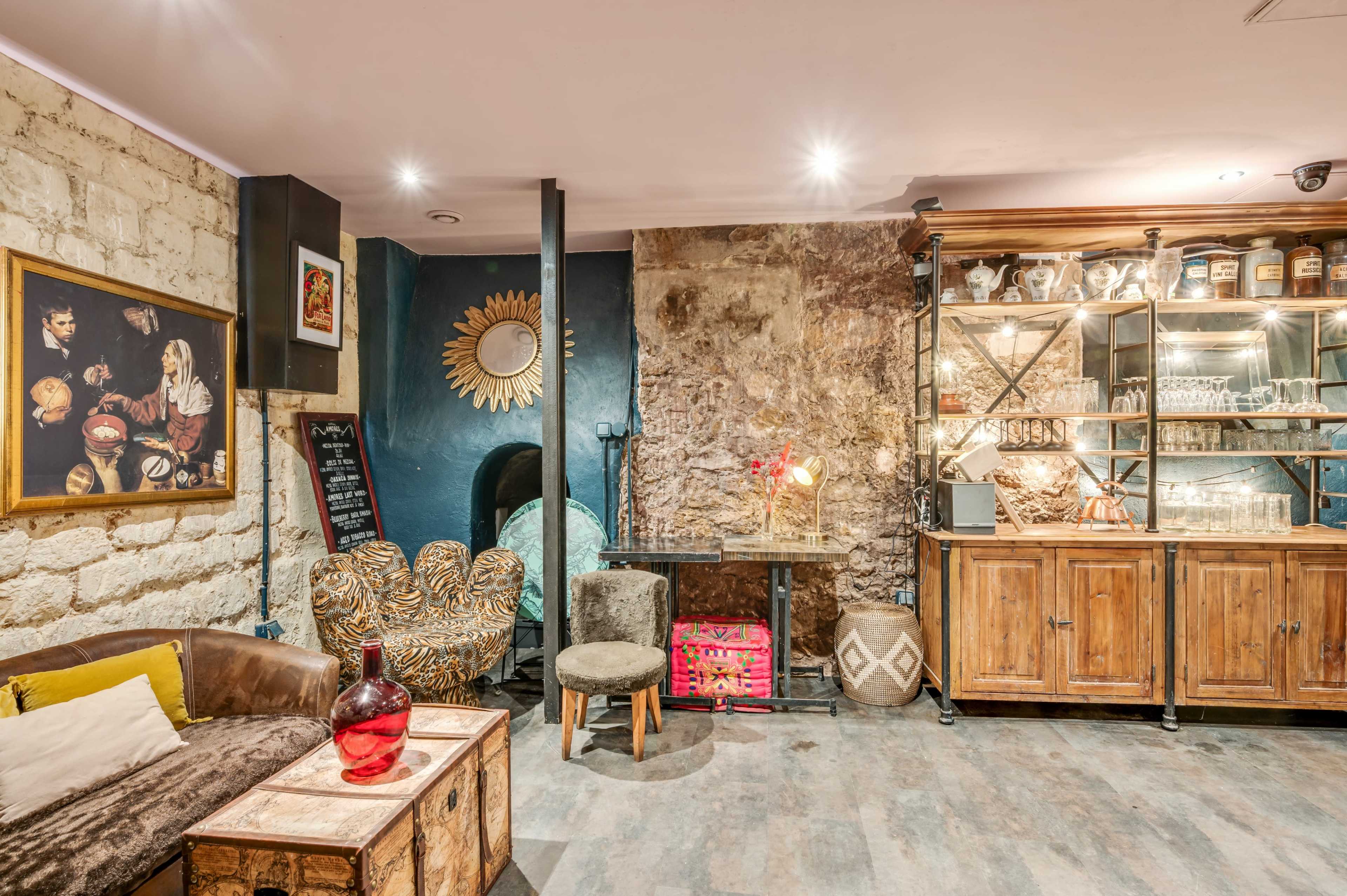 A cozy interior of a bar or lounge with a mix of vintage furniture, including a sofa, chairs, and a wooden cabinet filled with glassware, against a backdrop of exposed stone walls.