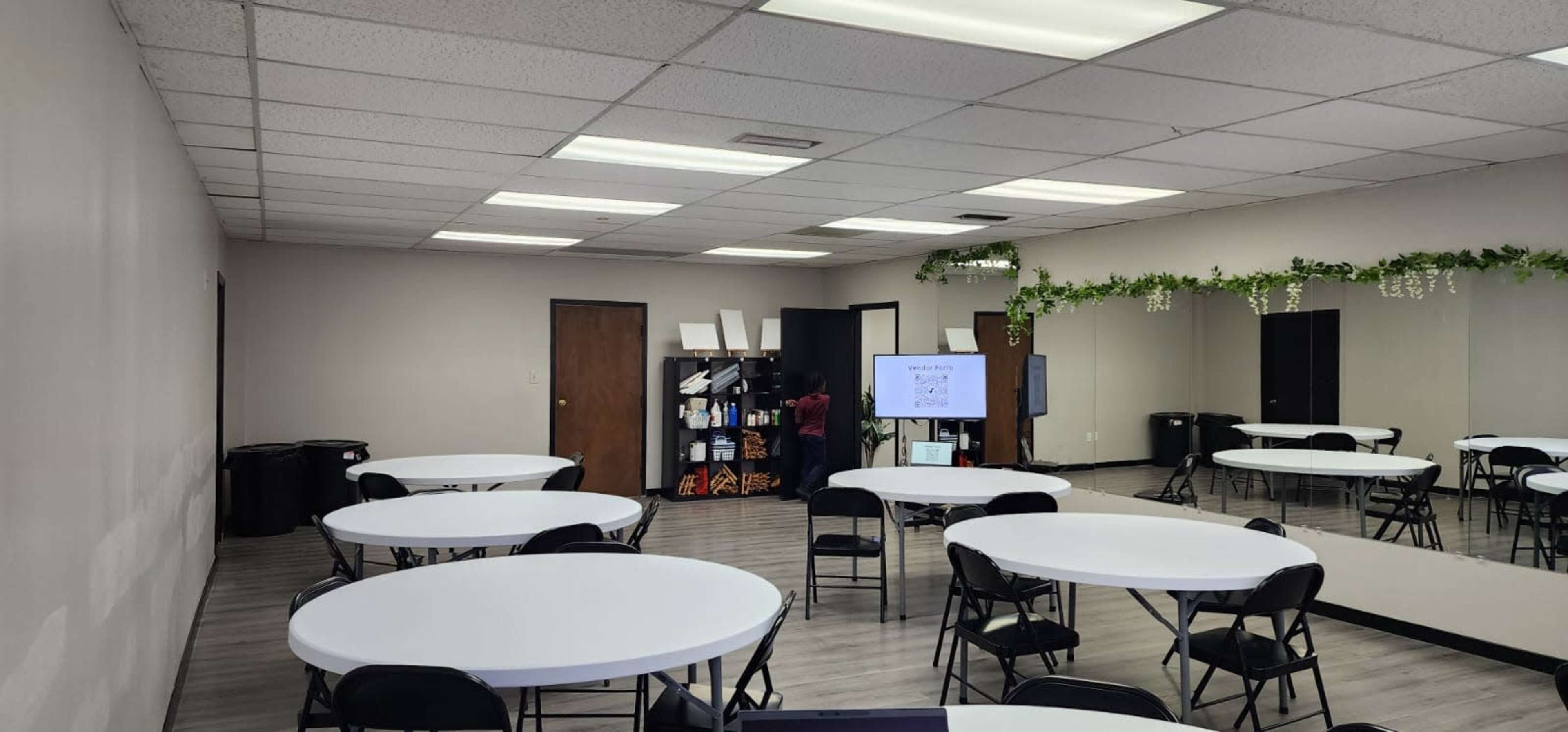 The image shows a spacious meeting room with several round tables and black chairs, along with a large screen and a mirrored wall.