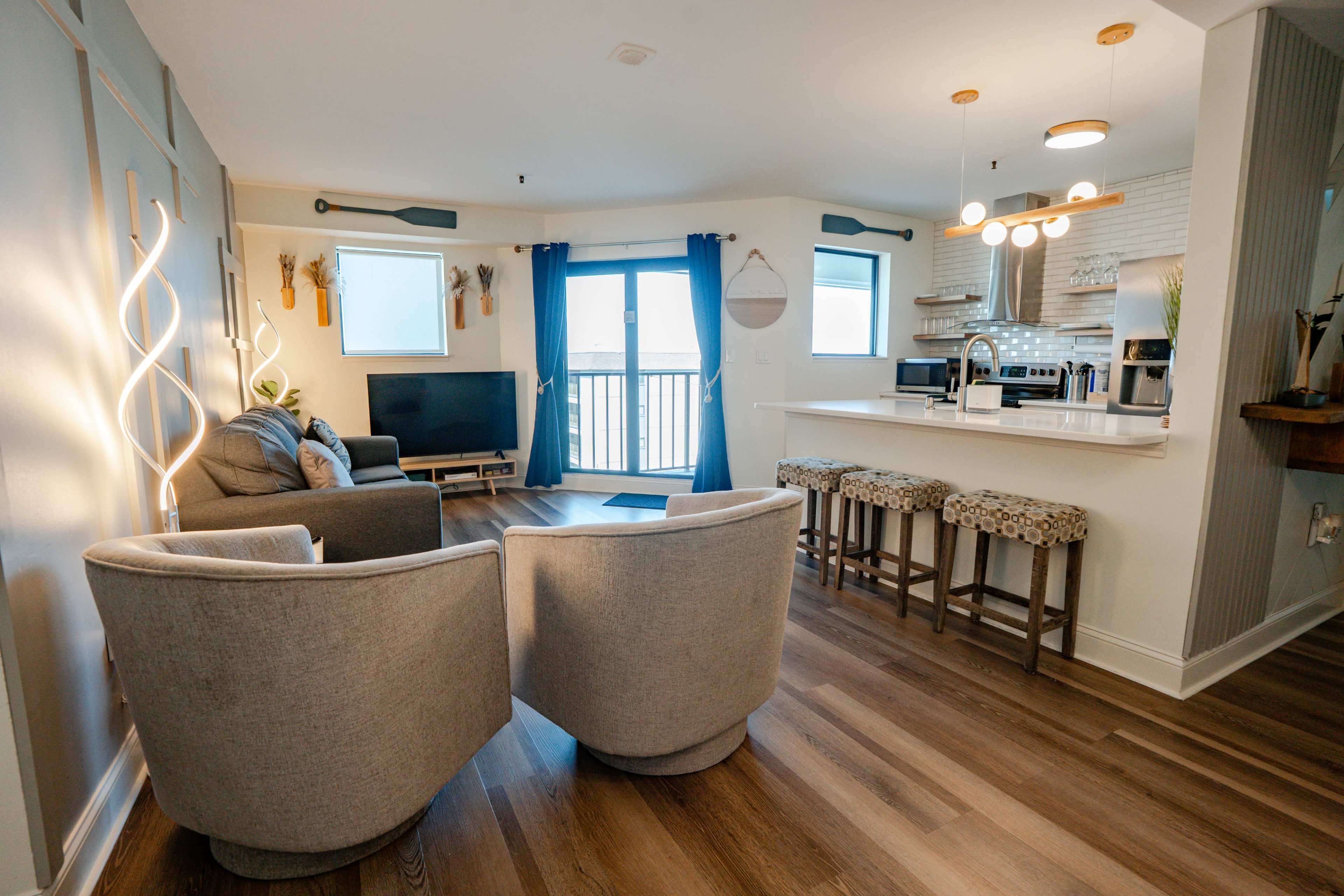 The image shows a modern living space with a cozy seating area, a television, and a kitchen bar with three stools near a balcony door.
