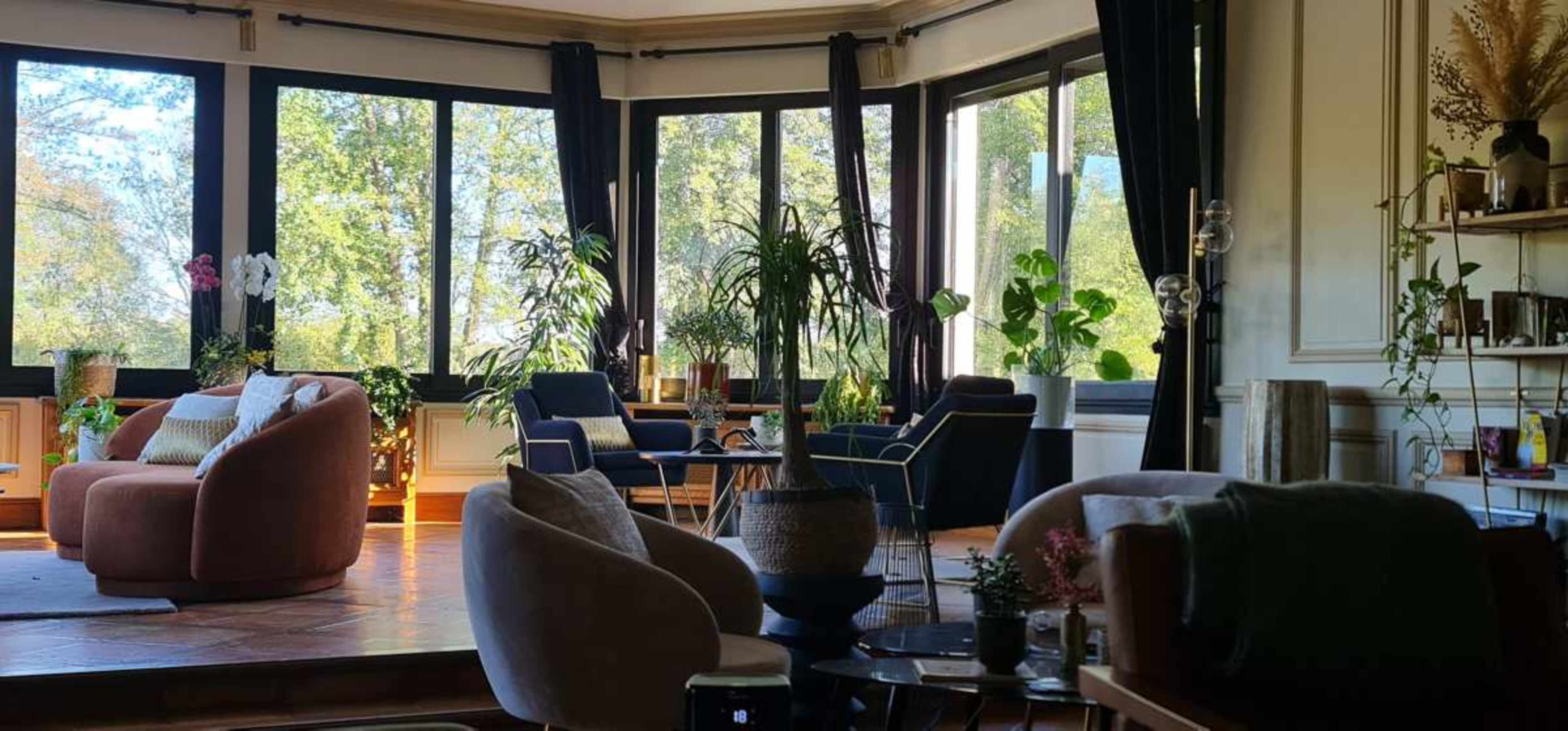 The image shows a bright, spacious room filled with green plants, large windows, and various cozy seating arrangements.