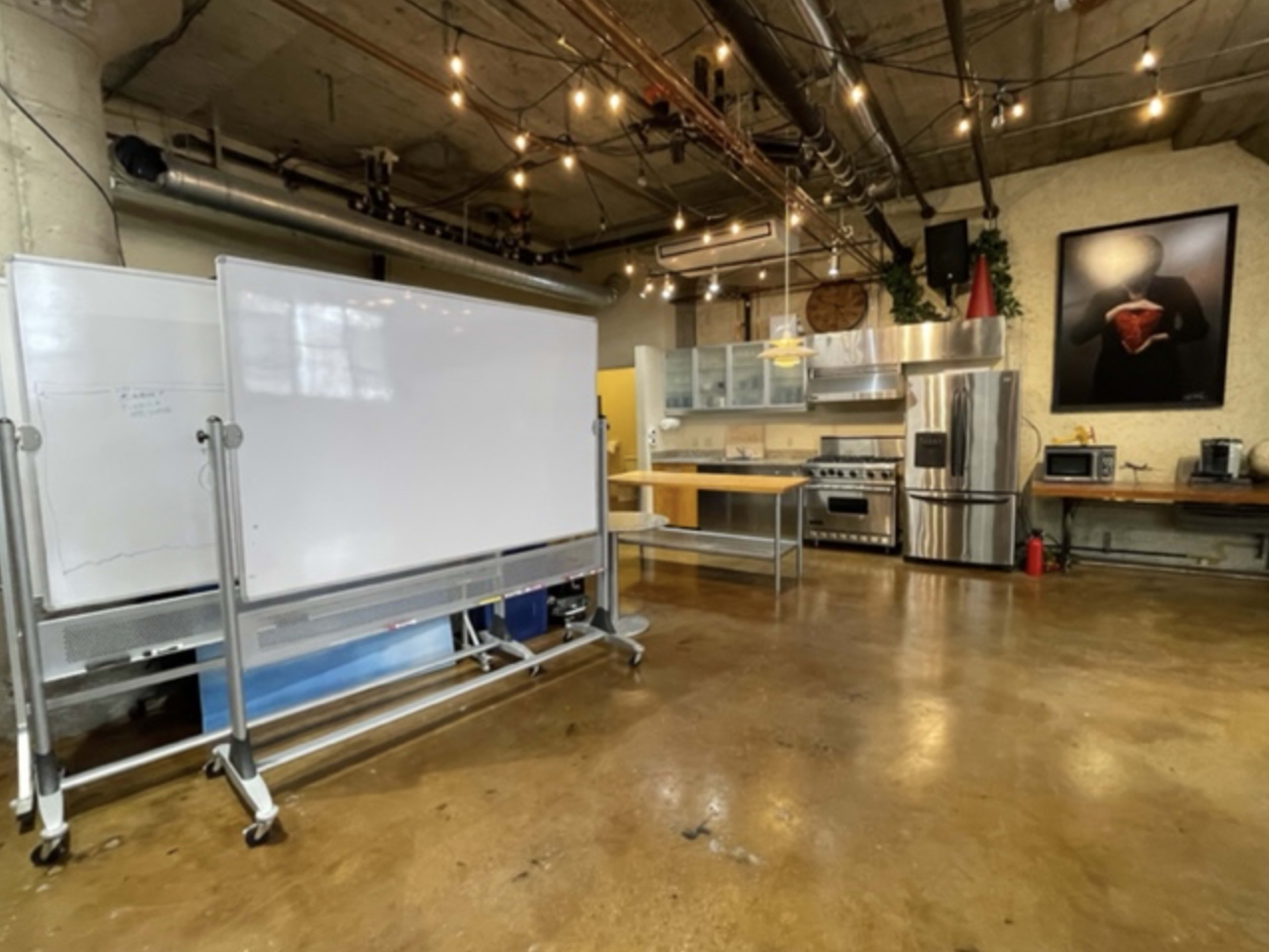 The image shows a spacious kitchen area in an industrial-style room with stainless steel appliances and a large whiteboard in the foreground.