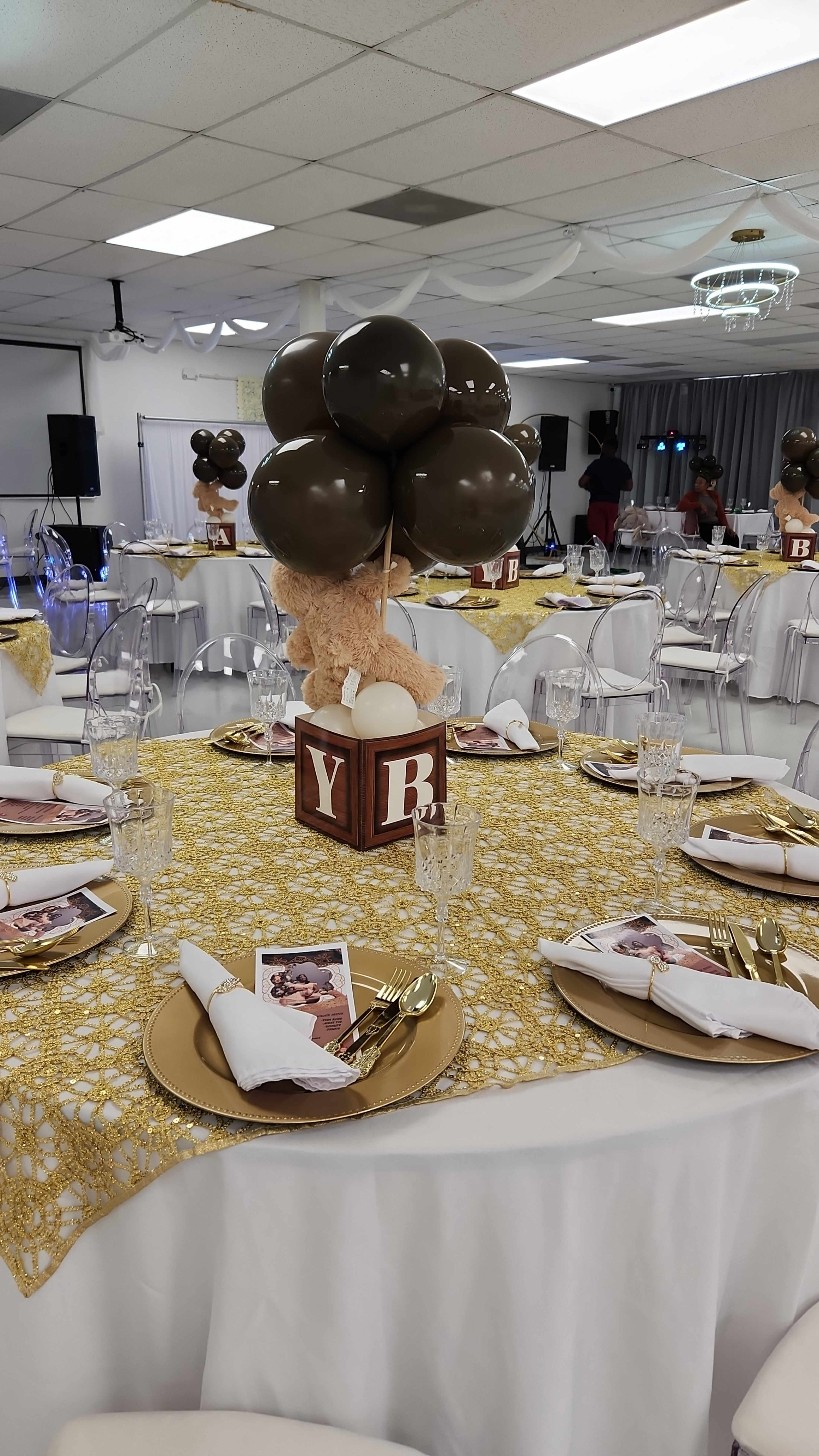 A decorative table is set for a celebration, featuring a gold tablecloth, clear chairs, and a centerpiece with balloons and a teddy bear.