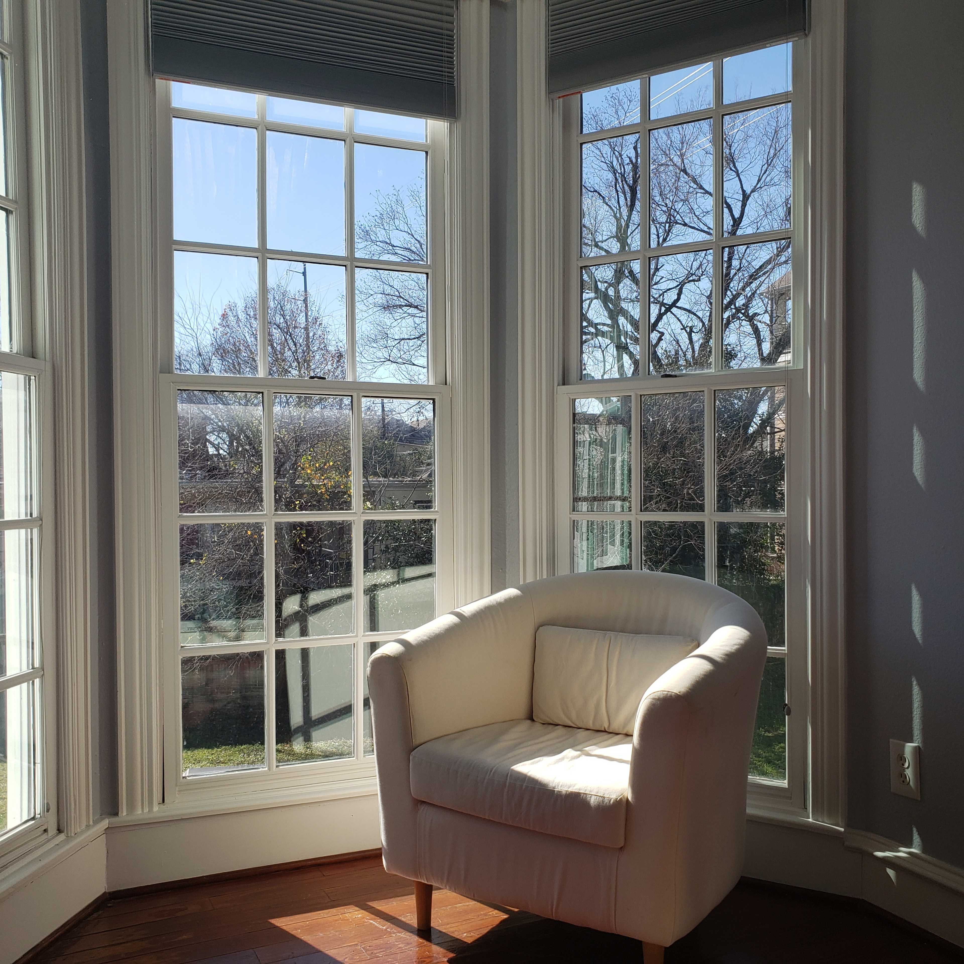 A white armchair is situated next to large, multi-paned windows that overlook a garden with bare trees.