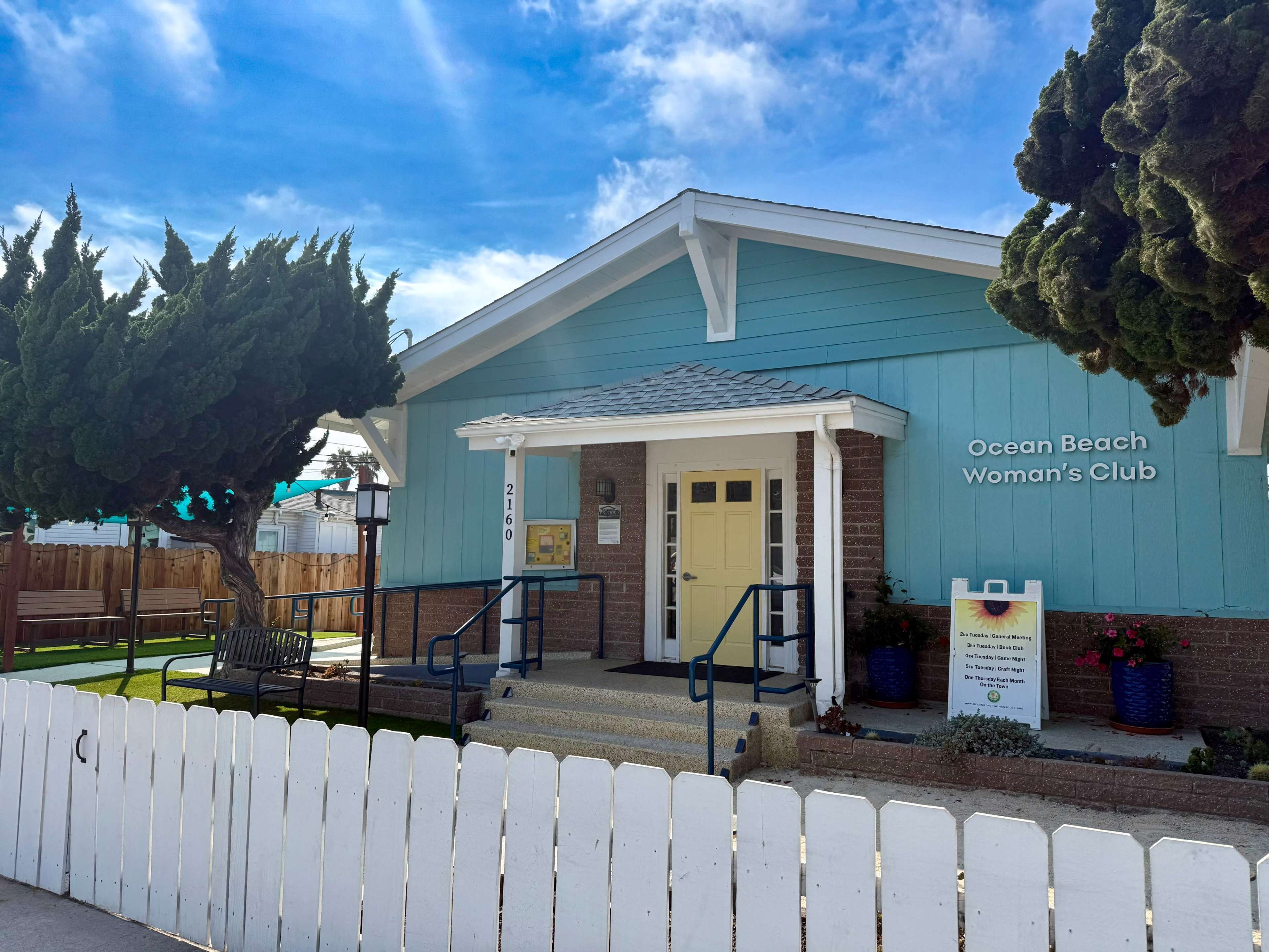 The image shows a light blue building with a sloped roof, labeled "Ocean Beach Woman's Club," surrounded by a white fence and green landscaping.