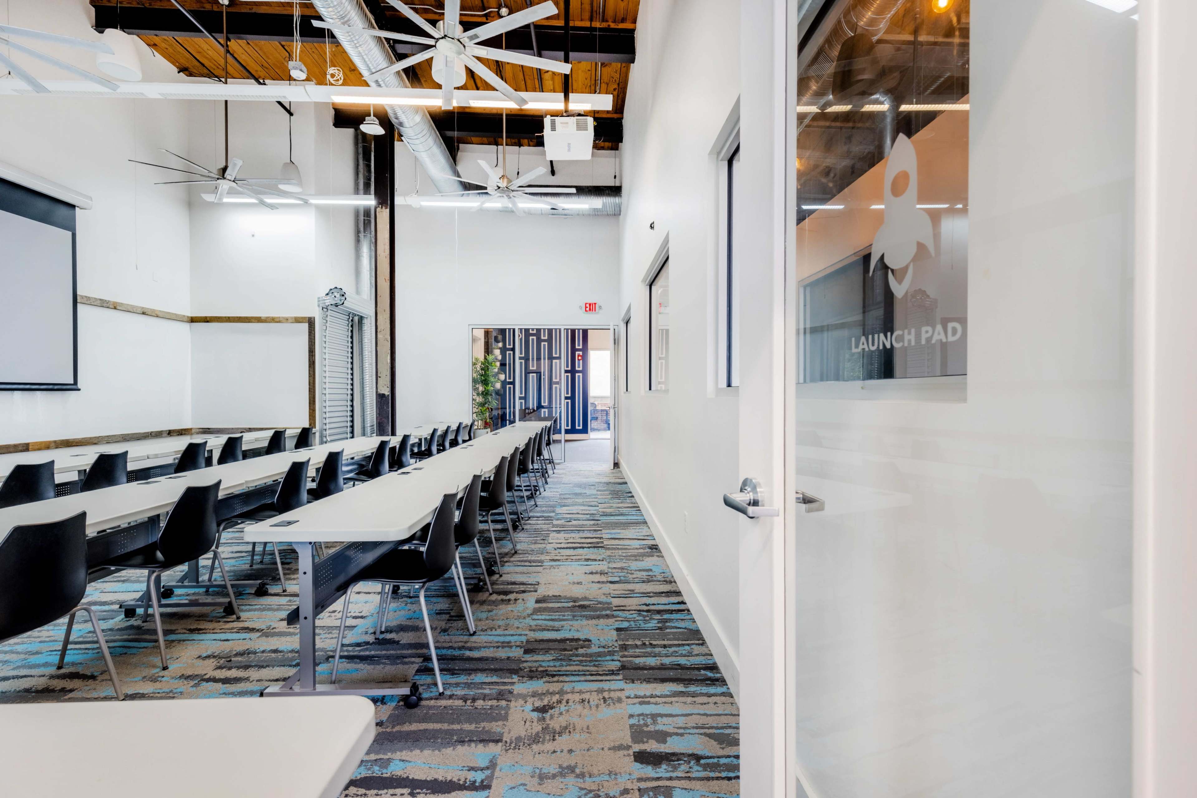 The image shows a modern, well-lit conference room with long tables arranged in a row and a screen on one wall.