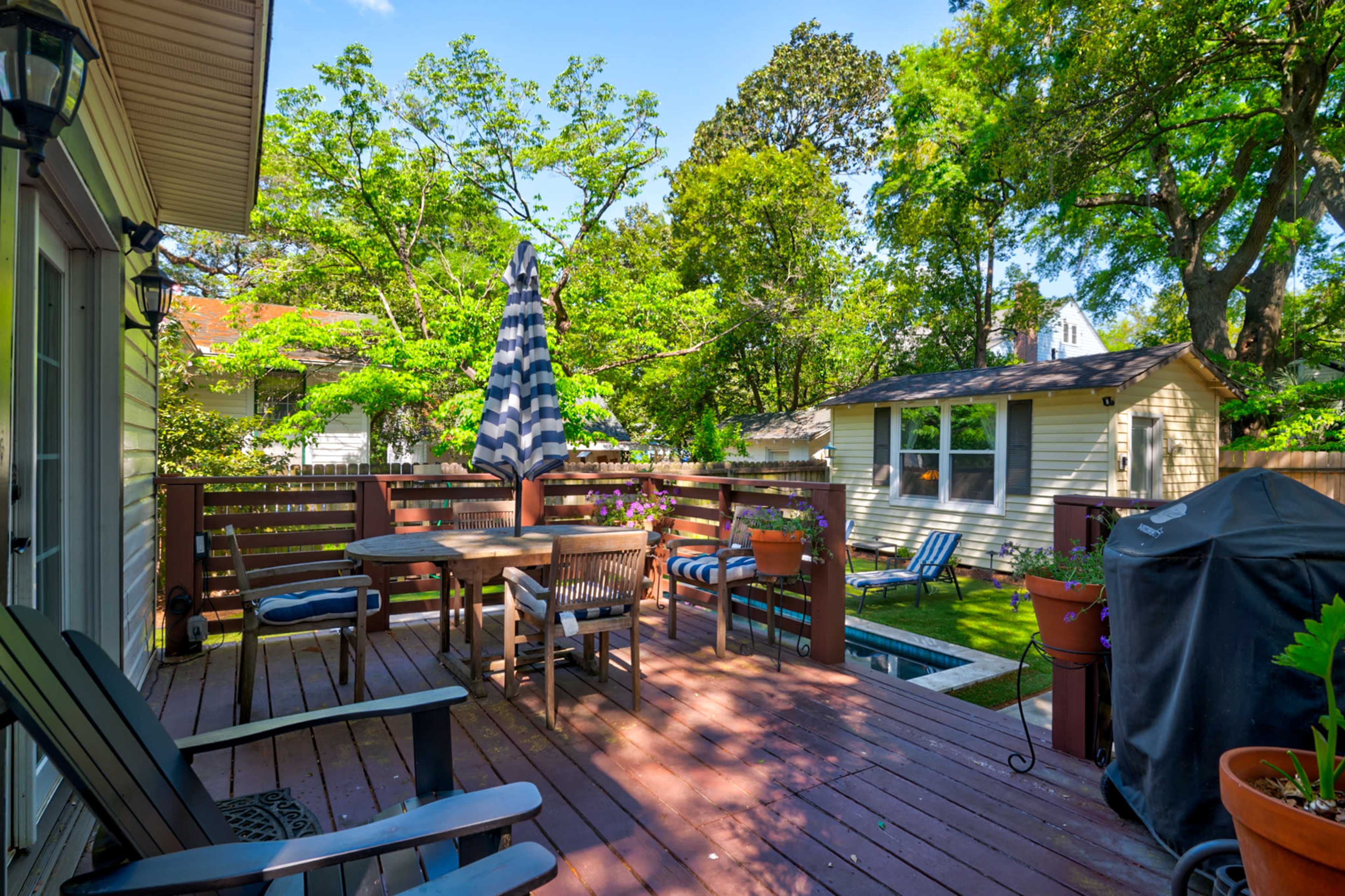 A wooden deck features a dining table with chairs, a striped umbrella, and a barbecue grill, surrounded by lush greenery and a nearby shed.