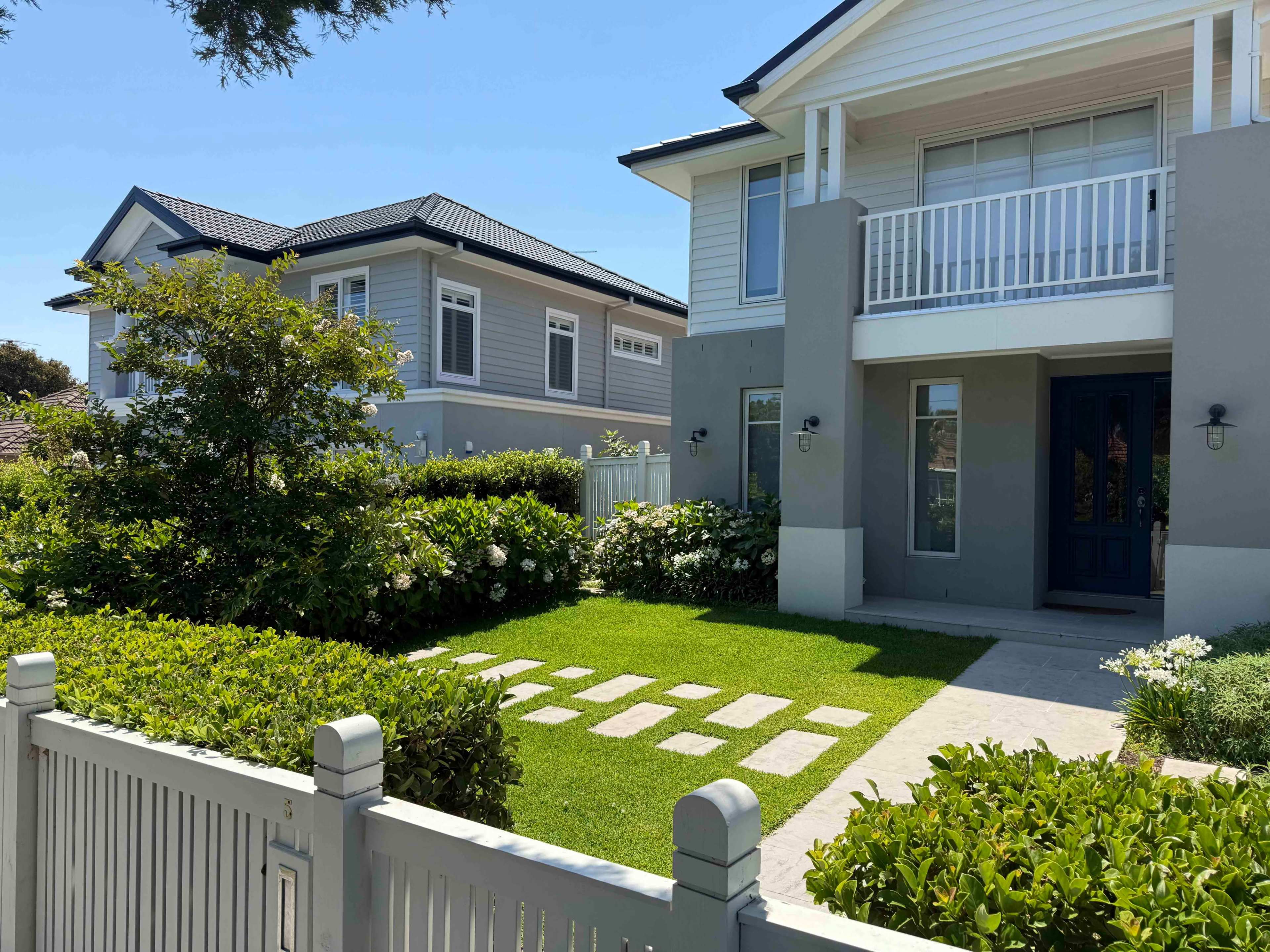 The image shows a well-maintained front yard with a pathway of stepping stones leading to a residential house, bordered by neat greenery and flowers.