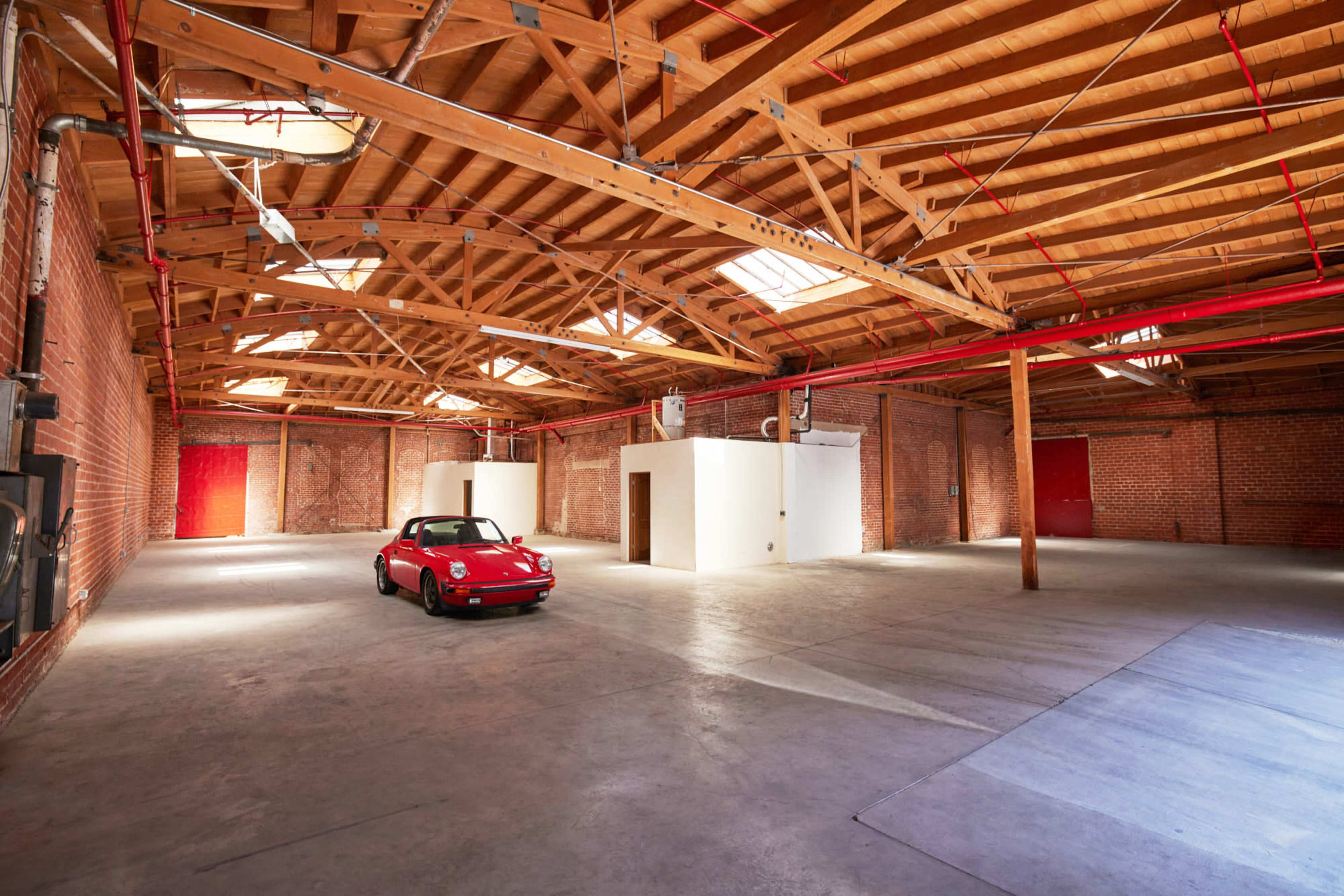 A red sports car is parked in a spacious warehouse with wooden beams and white walls.