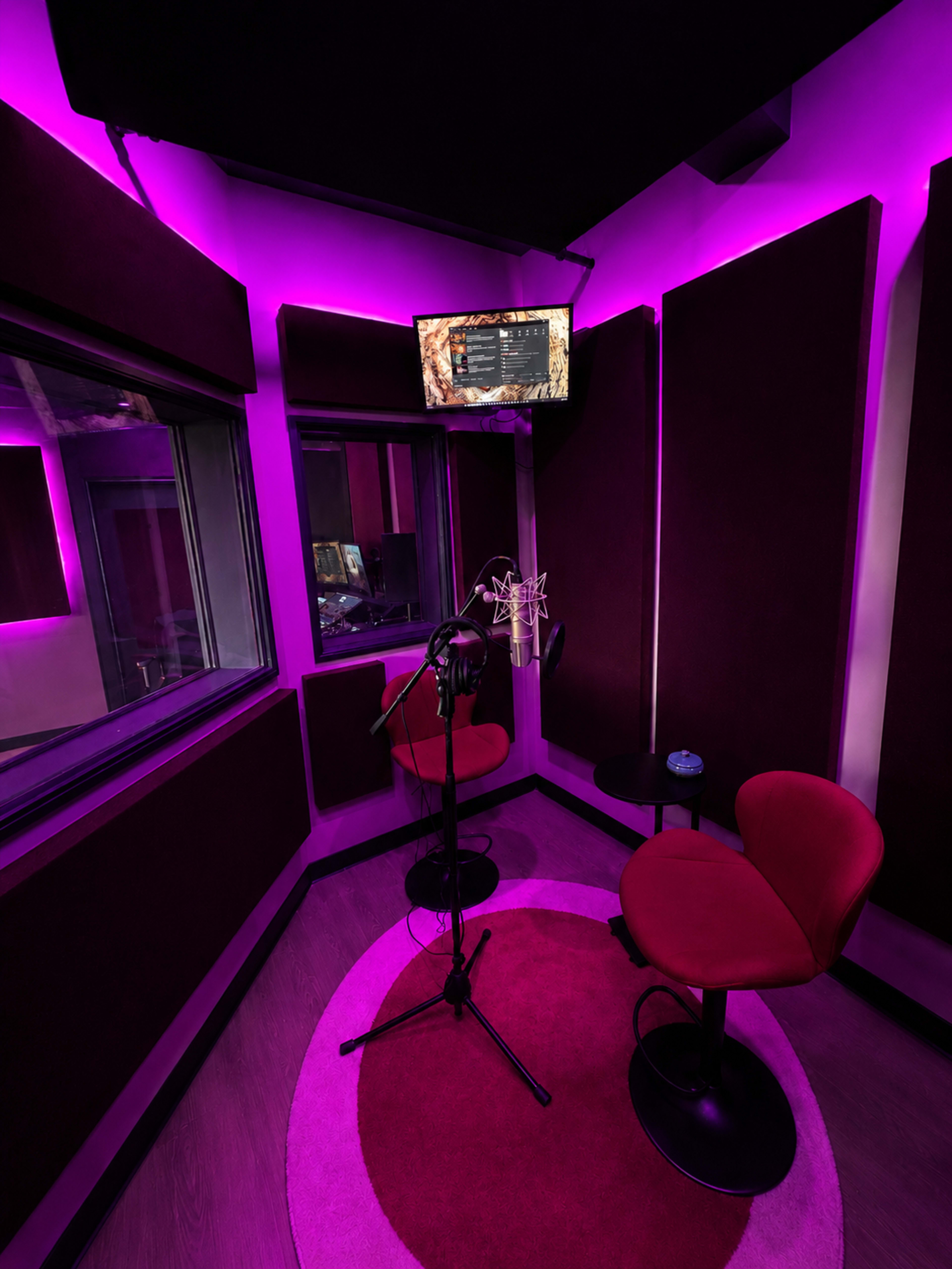 The image shows a recording studio with purple ambient lighting, featuring a microphone on a stand, two red chairs, and a round rug.