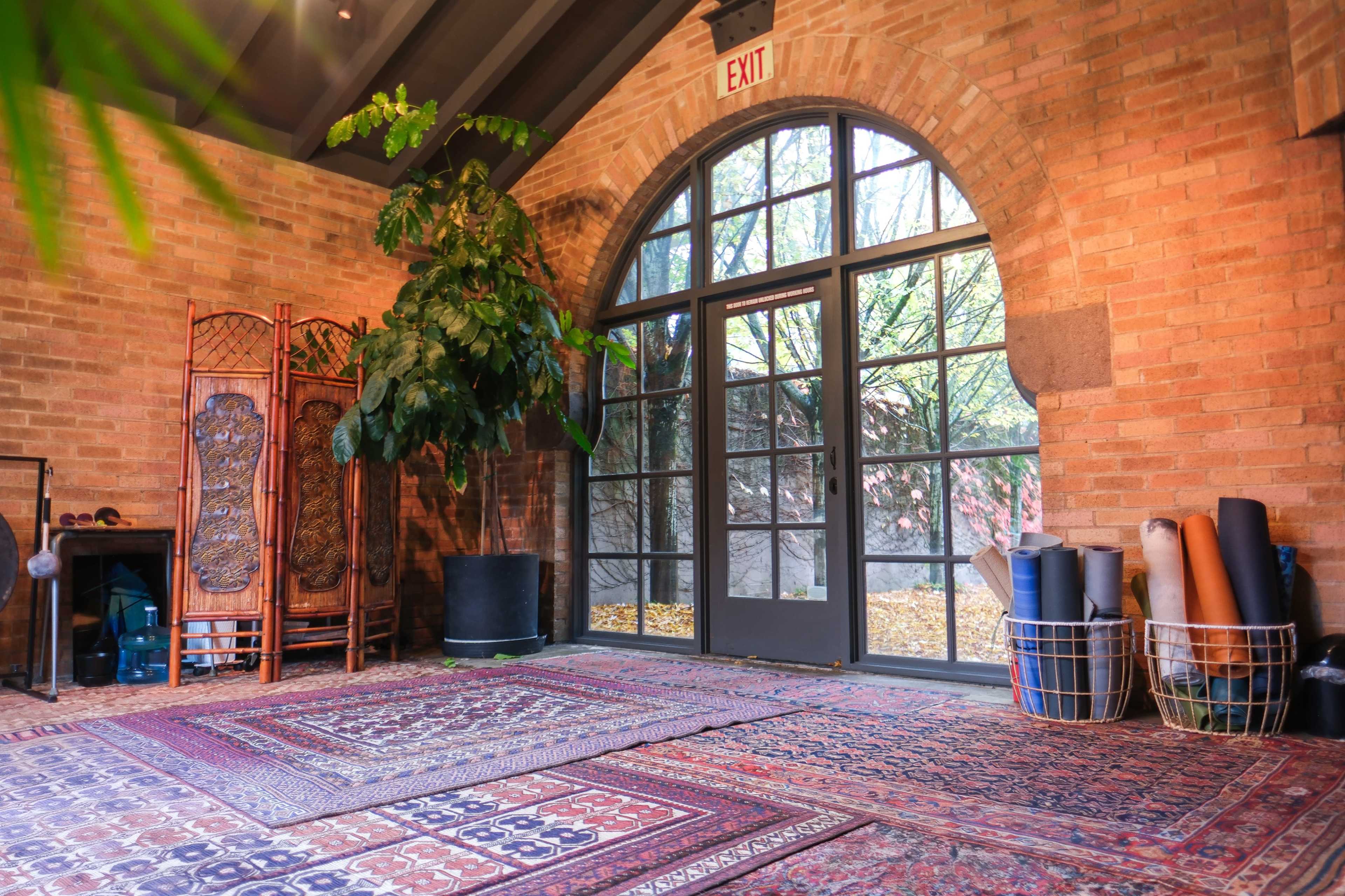 The image shows a spacious indoor area with a large window, wooden beams, brick walls, a decorative screen, and rolled yoga mats in baskets.