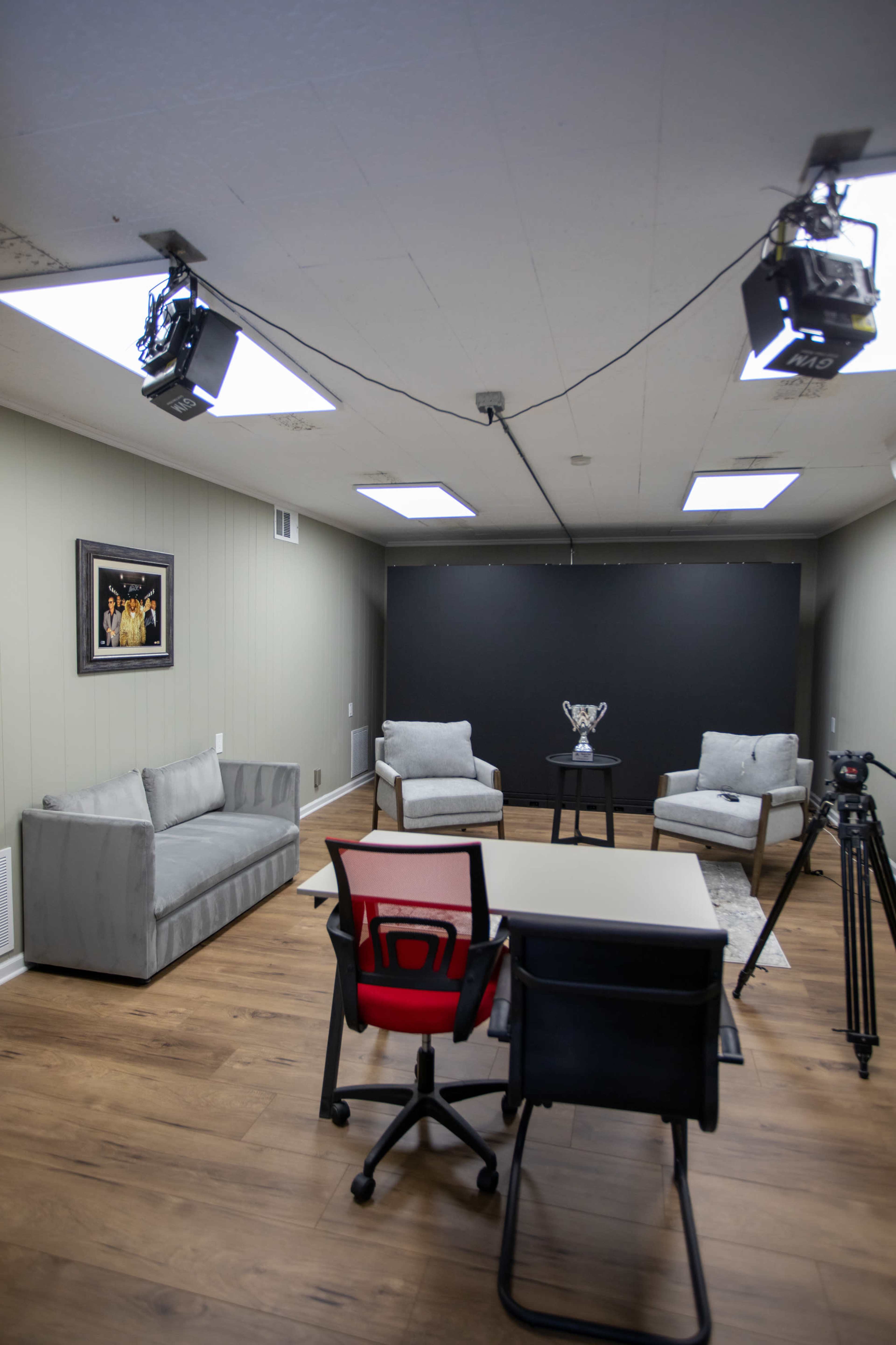The image shows a modern, well-lit recording studio with two armchairs and a couch facing a black backdrop, along with a table and a camera setup.
