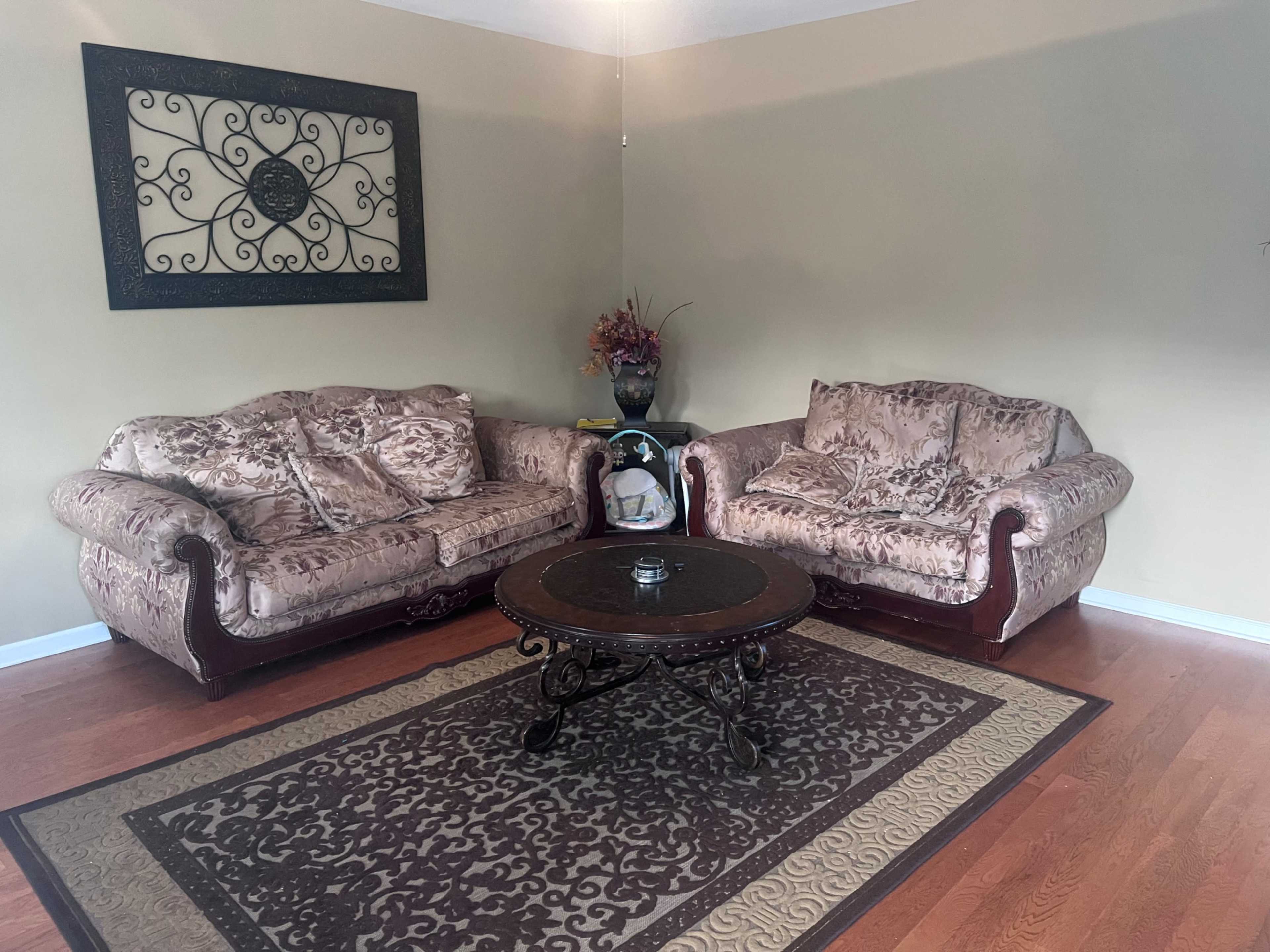 A living room with two ornate sofas facing each other, a round coffee table in the center, and a decorative wall hanging above.