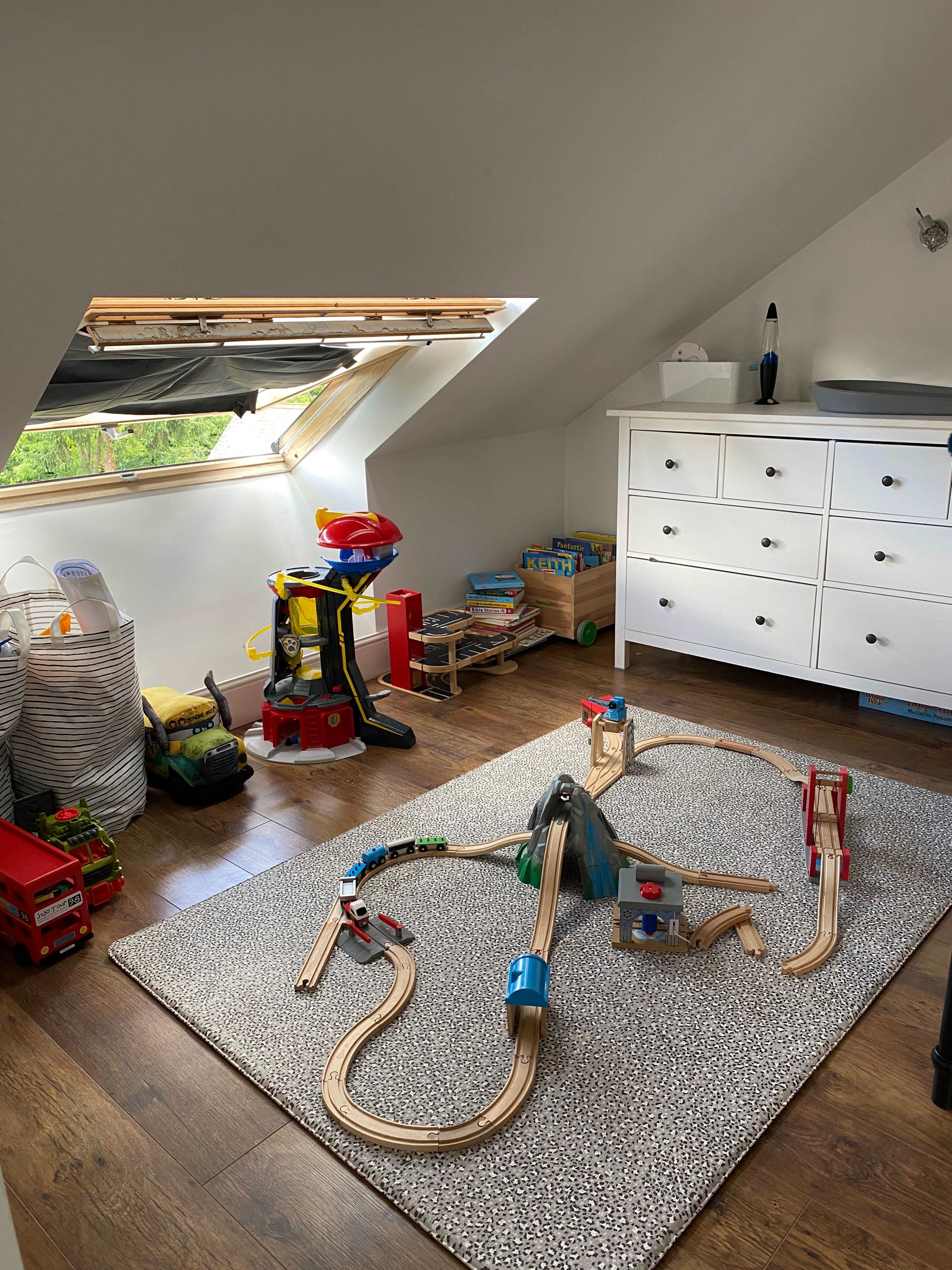 The room features a toy train set on a textured rug, with various toys and a dresser against the wall under a skylight.