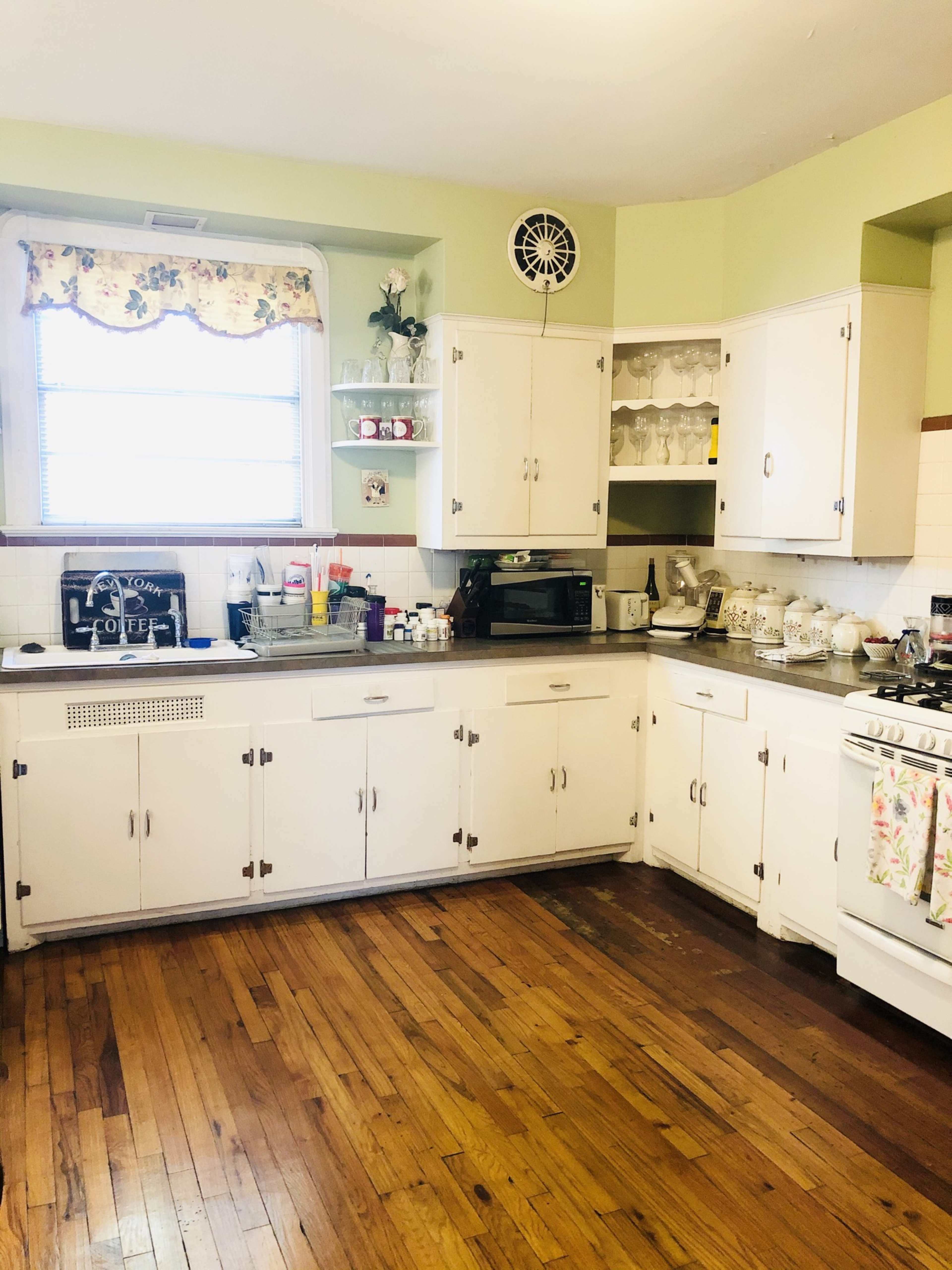 The image shows a kitchen with white cabinetry, a window adorned with floral curtains, and wood flooring.
